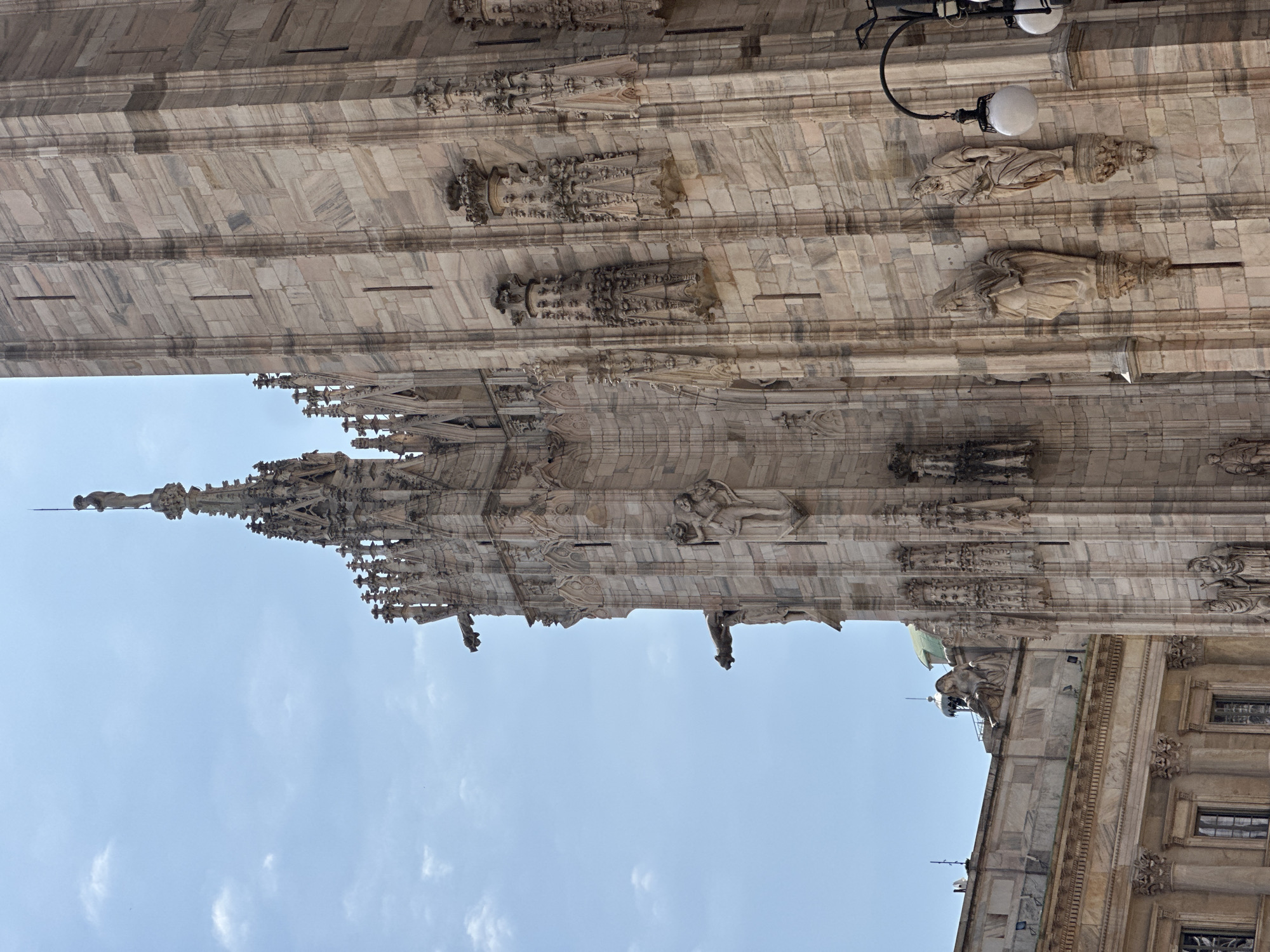 The image captures a close-up view of the Duomo di Milano, the cathedral of Milan, Italy. The facade is constructed of intricate, carved marble, with a dominant tone of light pink and beige. The architecture features Gothic spires, ornate sculptures of figures and gargoyles, and a towering wall of stone blocks. The sky above is a pale blue with scattered, wispy white clouds. To the left of the Duomo, a portion of another building with classical architectural elements, including columns and windows, is visible. A lamppost with two white globes is positioned on the right side of the frame, partially obscuring the cathedral's detail. The scene appears to be set outdoors during daylight.