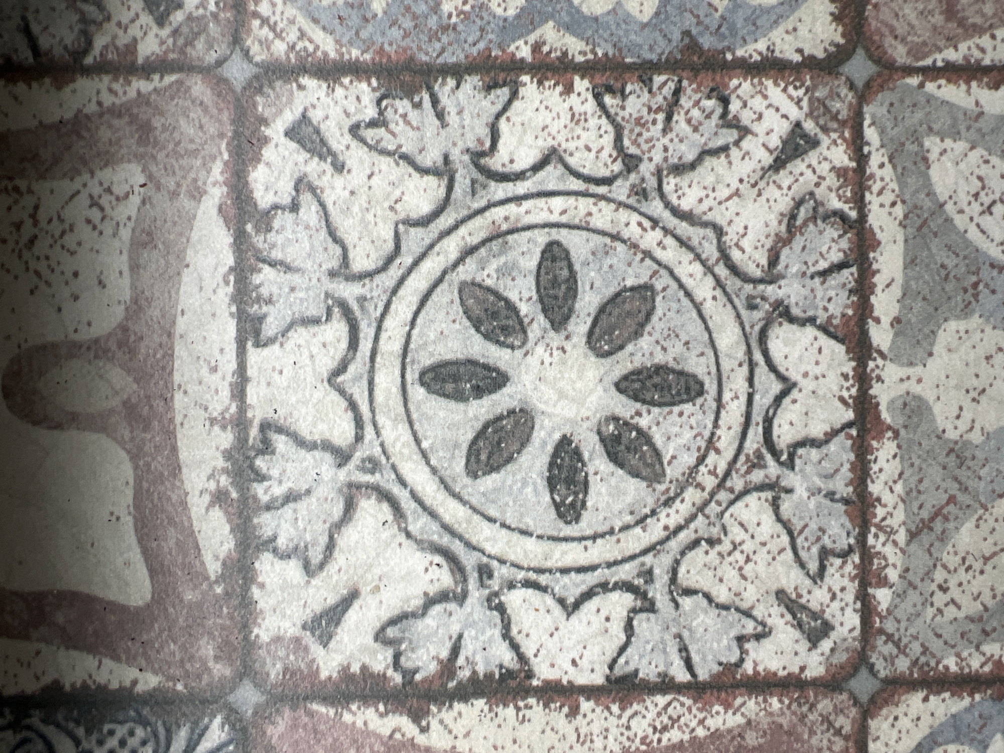A close-up photograph shows a patterned tile. The tile has a repeating floral design with a central circular motif containing six petal-shaped elements. An outer ring of leaf-like shapes surrounds the central circle. The tile is part of a larger surface composed of similar tiles, visible at the edges of the frame. The colors are muted, featuring shades of grey, white, and a reddish-brown. There is a weathered or distressed appearance, with a speckled texture and slight discoloration. The location is Kommunarka, Russia.