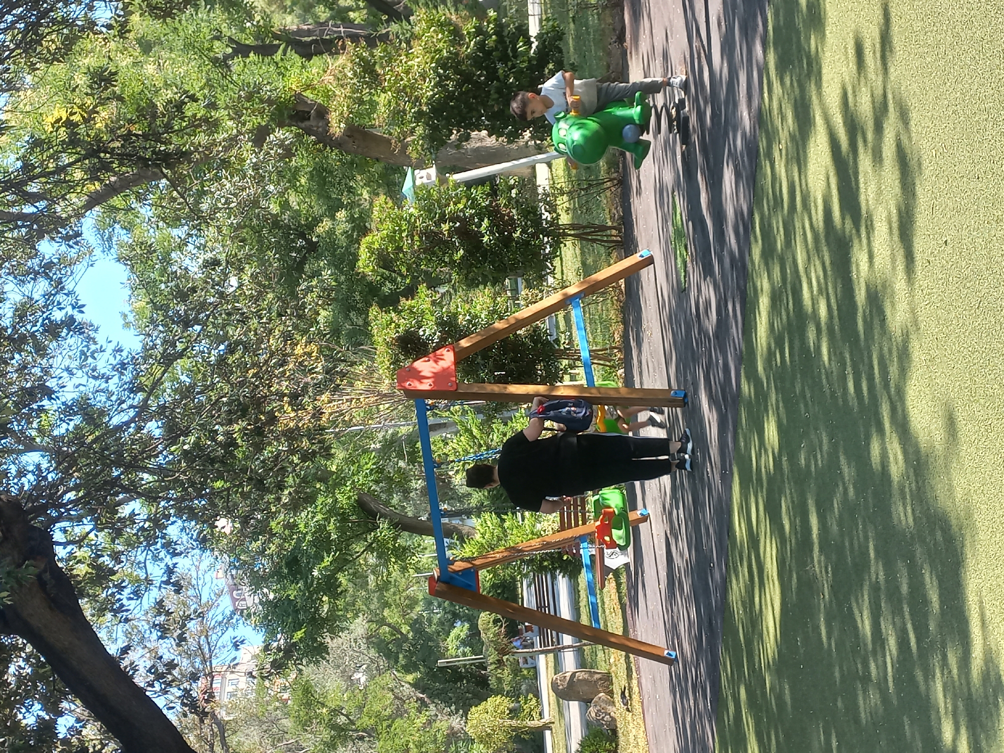 The image depicts a park scene with a playground. In the foreground, a child is playing on a green spring rider shaped like a hippopotamus. To the left, an adult stands near a swing set. The playground is situated on a paved area with patches of artificial turf. In the background, lush green trees and bushes create a shaded environment. A building is visible through the trees in the distance. The overall impression is one of a sunny day in a well-maintained urban park. The specific location of Baku, Azerbaijan is not visually verifiable from the image itself.