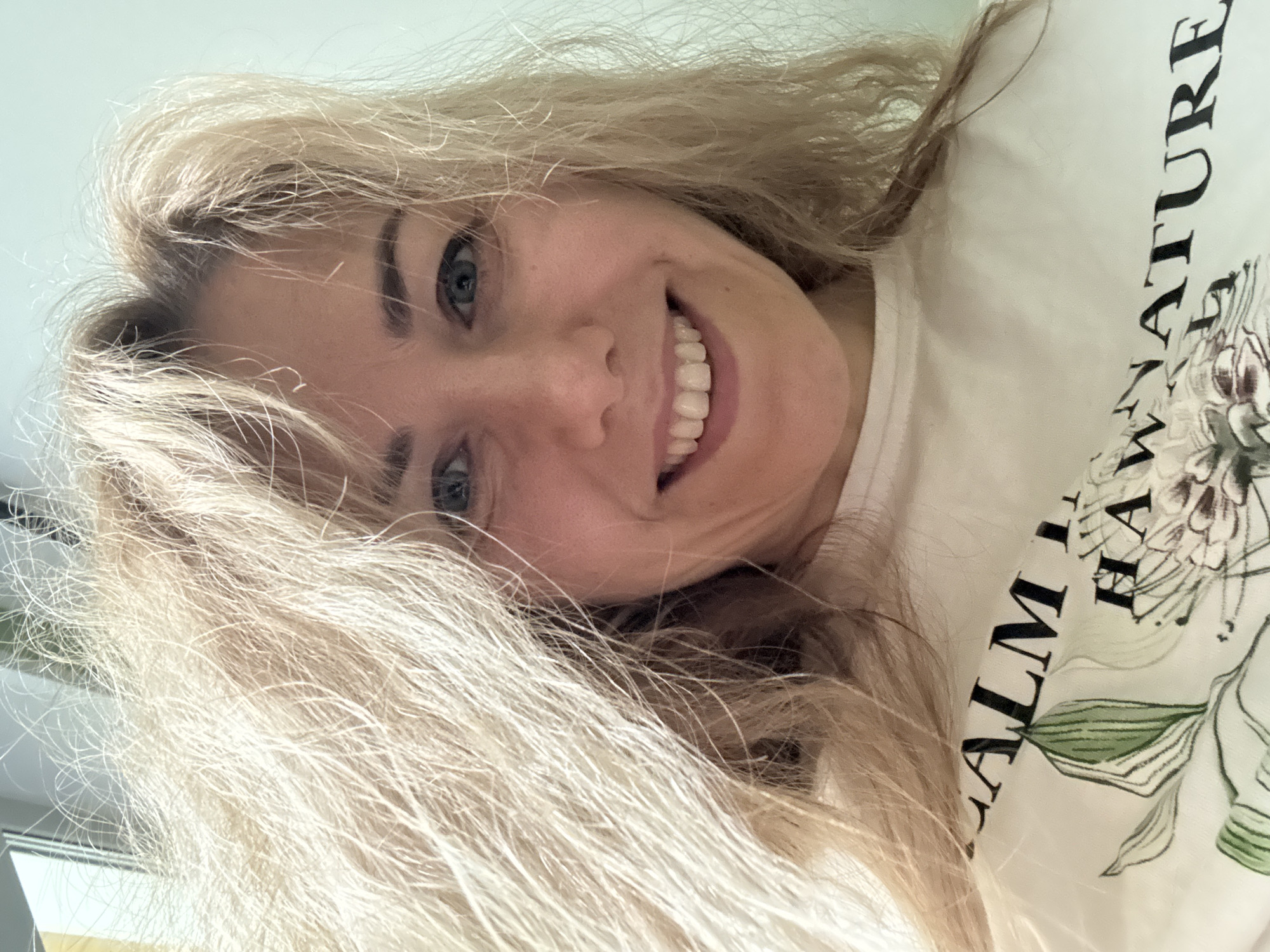 A blonde woman is smiling at the camera in a close-up shot. She is wearing a white t-shirt with a floral design and the text "CALM NATURE". The photograph appears to be taken indoors. The location is not discernible.