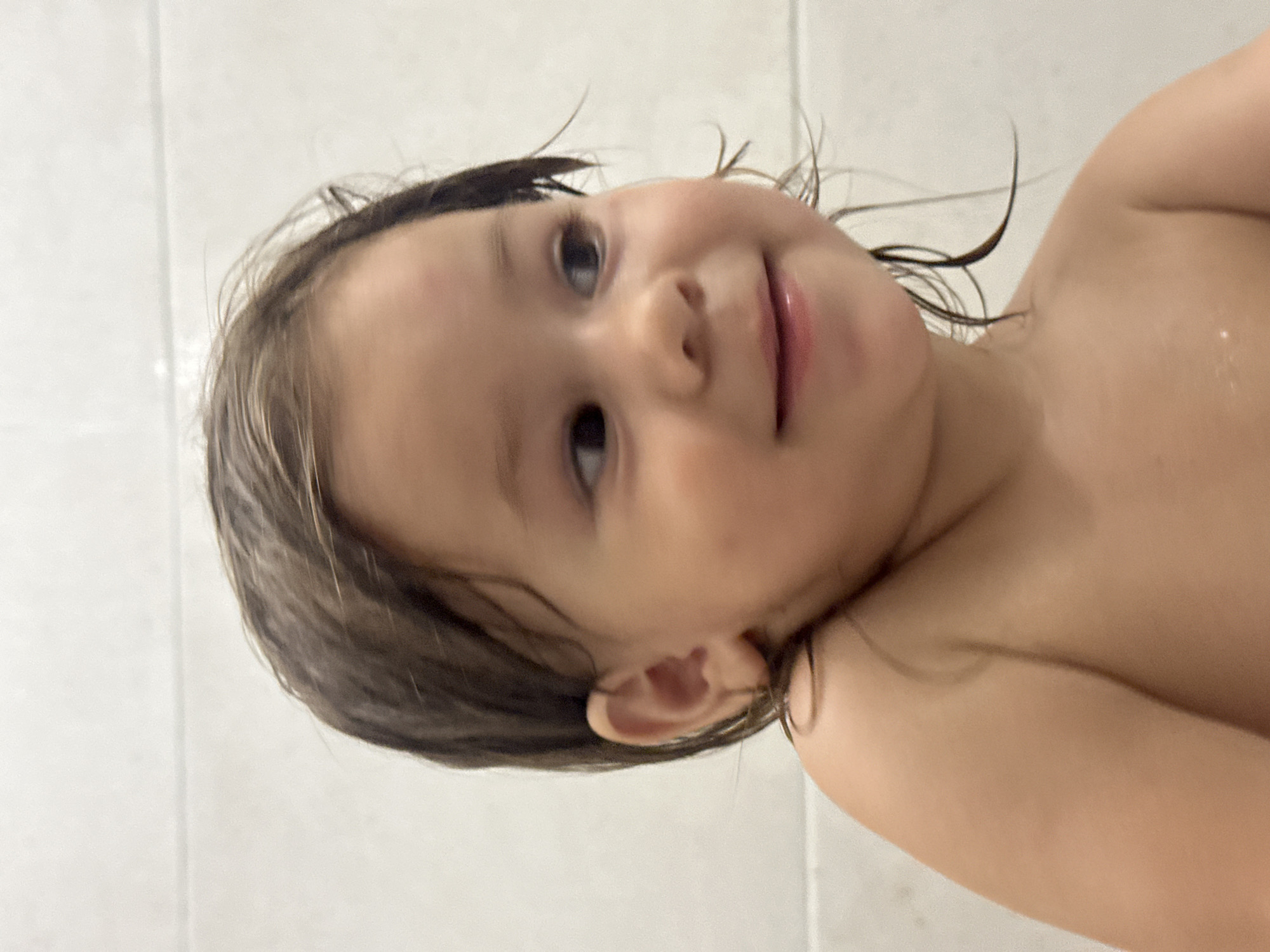A close-up, vertical, medium shot shows a young child's face from a slightly low angle. The child, appearing to be a toddler, has dark, wet hair that is styled in loose strands around their head and face. They are looking upwards and to the left with a happy and playful expression, their mouth slightly open in a smile. The lighting is soft, suggesting an indoor setting. The background is a plain, light-colored surface, possibly tiles, indicating a bathroom or shower area. The child's skin is smooth and appears slightly flushed, which could be from a bath or being warm. A part of the child's shoulder and arm is visible on the right side of the frame, also appearing damp. The overall scene conveys a sense of innocent joy and a moment of relaxed play, likely during bath time. No text is visible in the image.