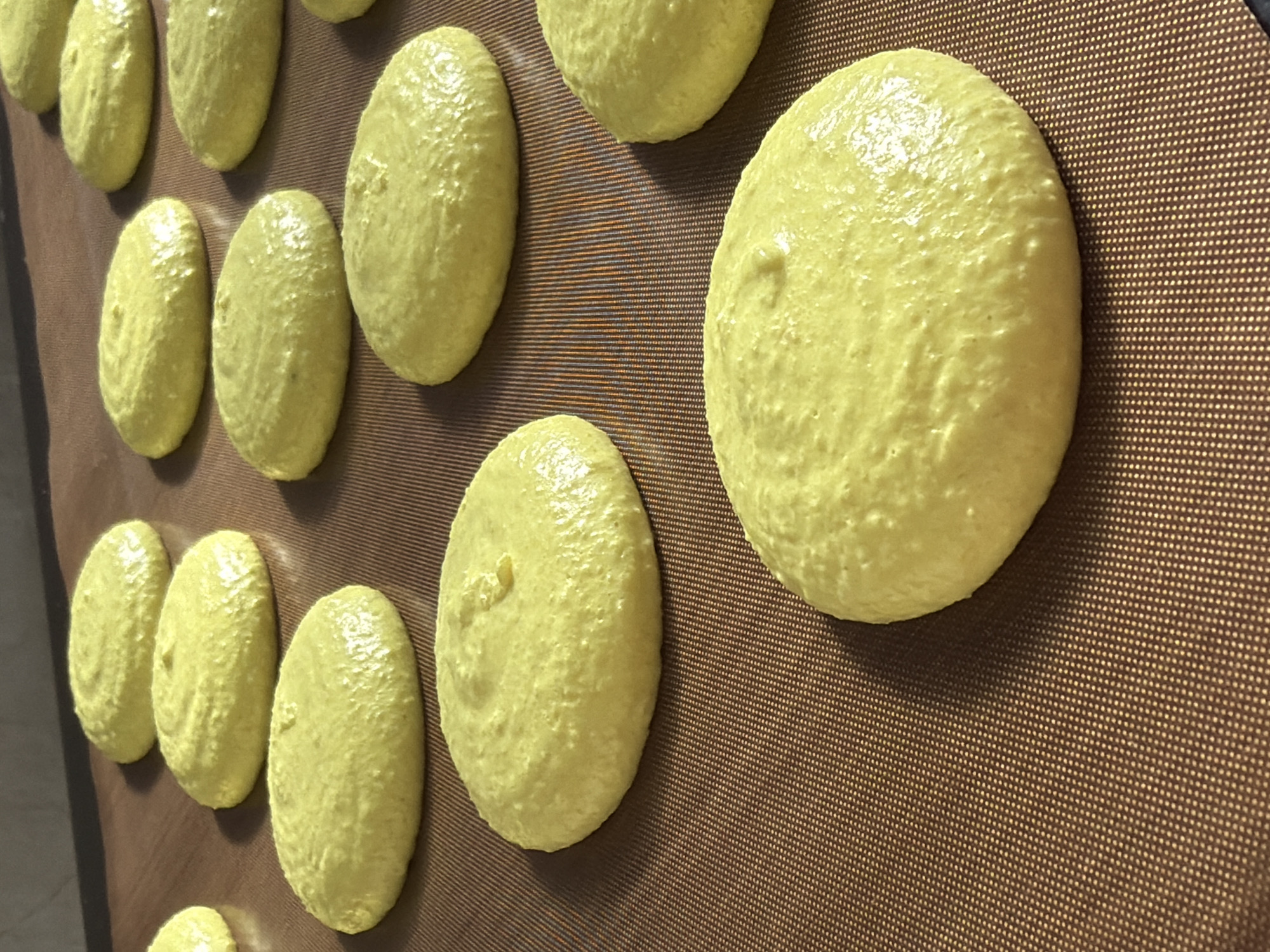 A baking sheet filled with yellow macarons rests on a brown silicone baking mat. The macarons are arranged in rows and appear uncooked. The setting is indoors. There is no indication of the location being Tel-Aviv, Israel.