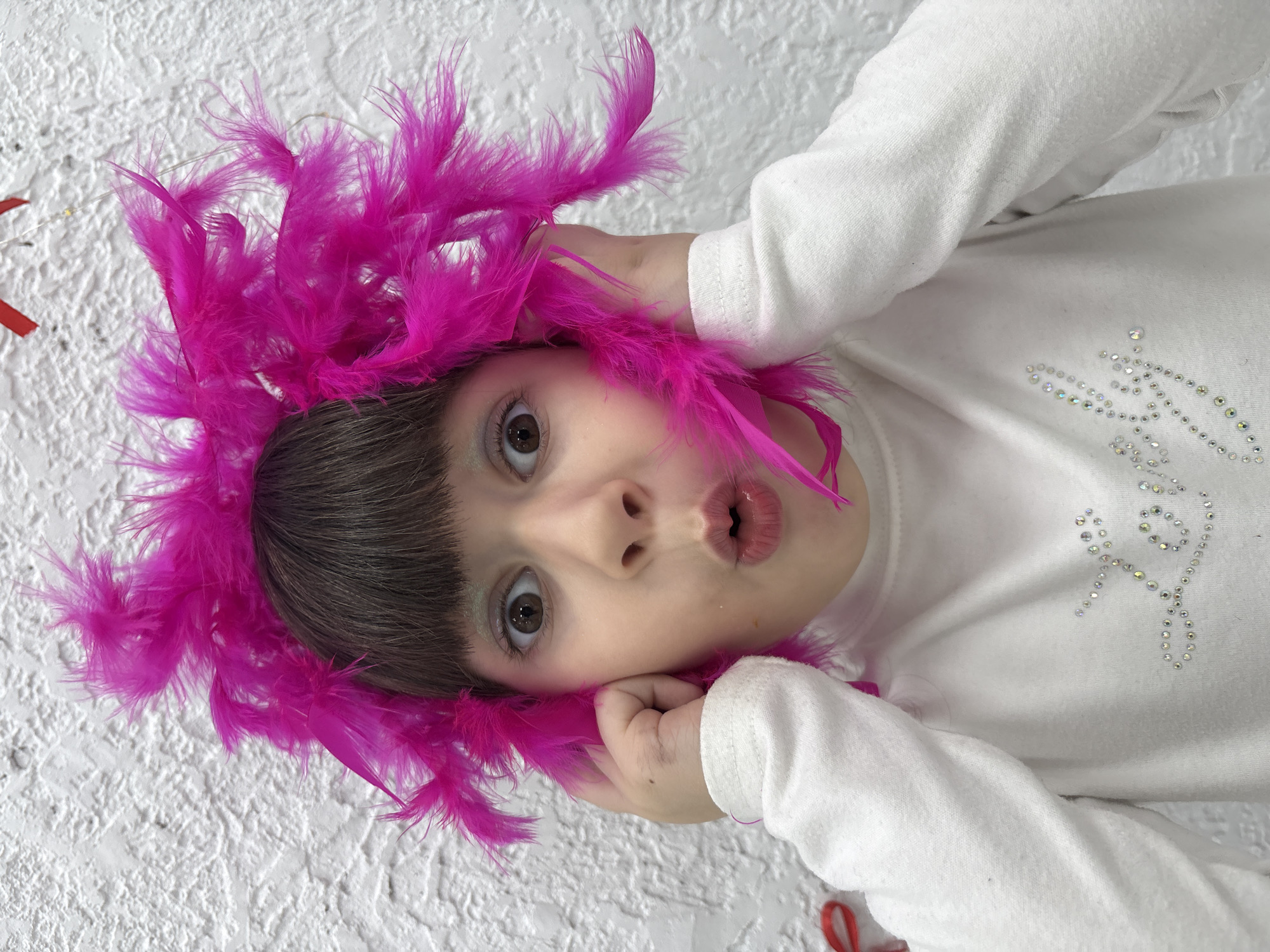 A young child is centered in the frame, looking up with a surprised expression and pursed lips. She is wearing a white, long-sleeved shirt adorned with a decorative pattern of rhinestones on the chest. Her hair is dark brown and straight. Around her head, there is a vibrant pink feather boa. The child is holding parts of the boa near her ears, framing her face.
The background appears to be a textured white wall. The image is oriented vertically. Given the prompt’s specifications, no further information about the location is discernible from the visual data provided.