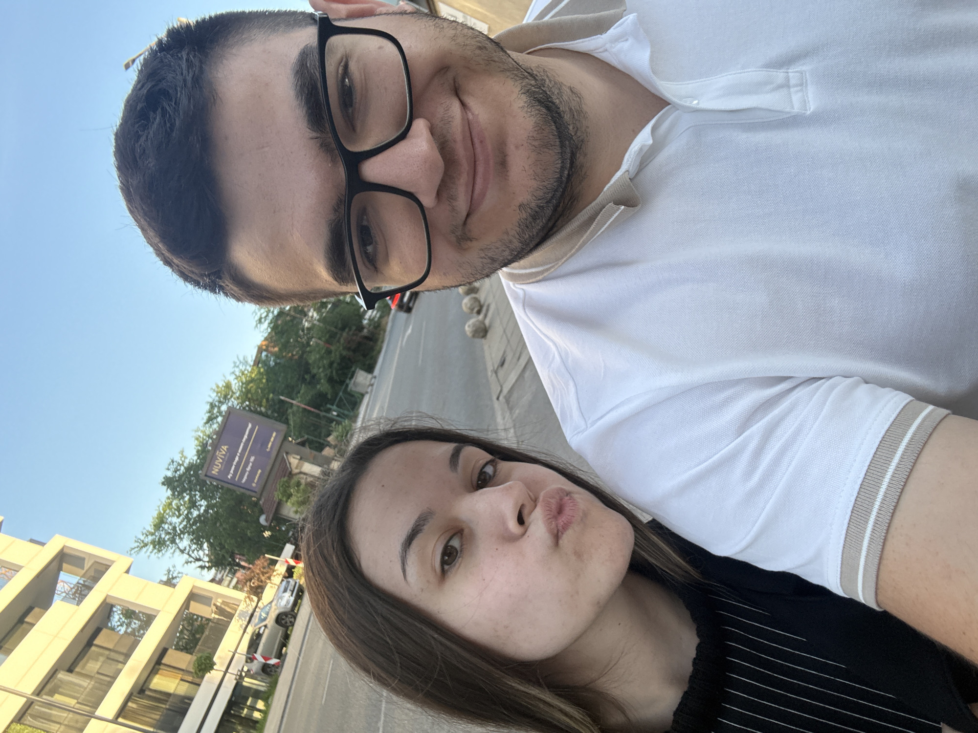 Two individuals are taking a selfie. The person in the foreground is a woman with brown hair and is pouting her lips. Behind her is a man wearing glasses and a white polo shirt. The background includes a street, buildings, and signage. The sign in the background indicates the location is near the Nuviva Clinic. Given the signage and architecture, the location is likely in the city of Sofia, Bulgaria.