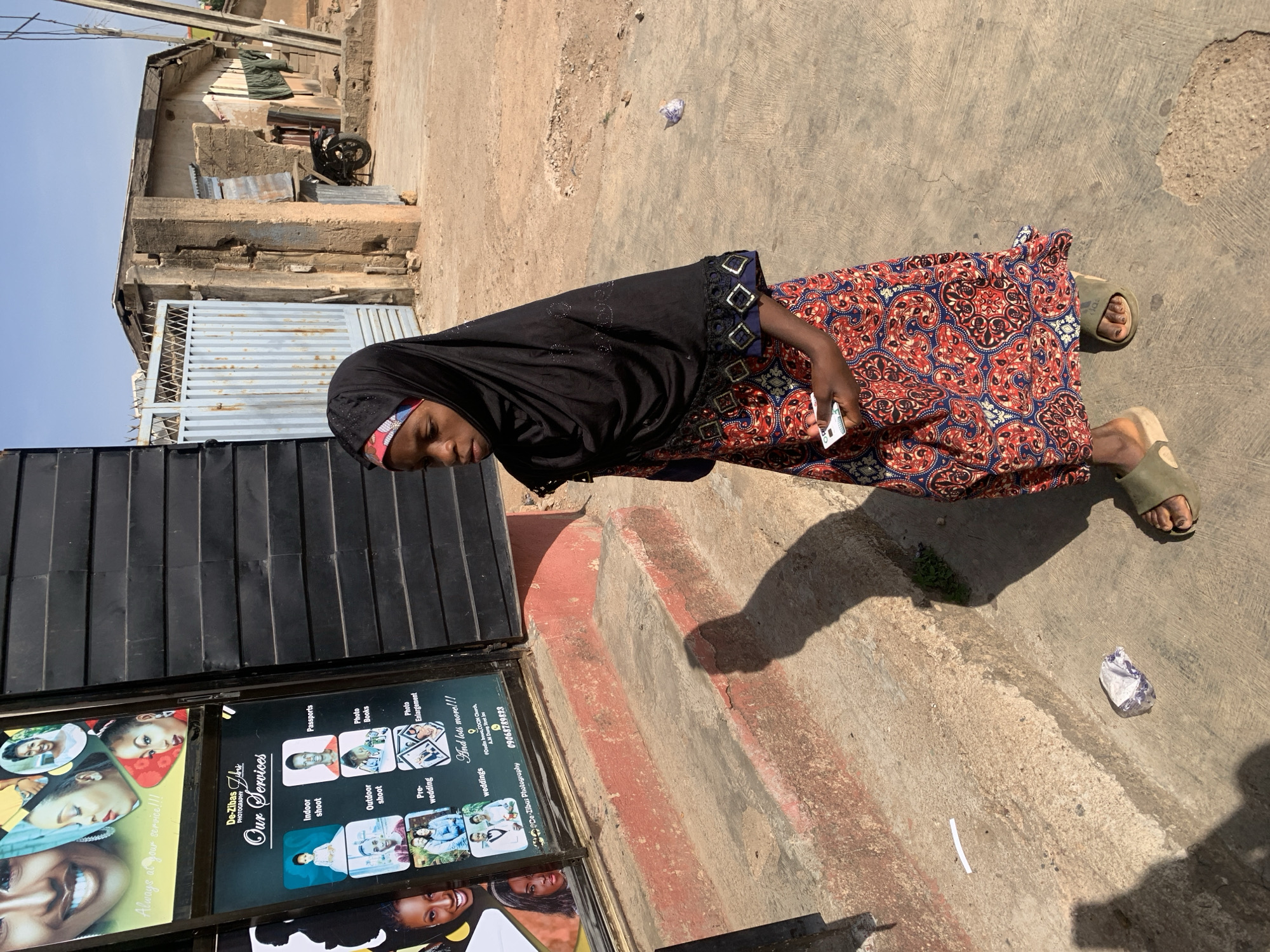 A woman wearing a black head covering and a patterned dress stands on a concrete sidewalk in Jos, Nigeria. She holds a white object in her hand. Next to her is a sign advertising photography services, including indoor and outdoor shoots, passports, photo books, pre-wedding, and weddings. The address on the sign is near COCIN Church on A.M. Dung Street. Behind the woman is a building with a corrugated metal gate. The weather is sunny. There is trash on the sidewalk.