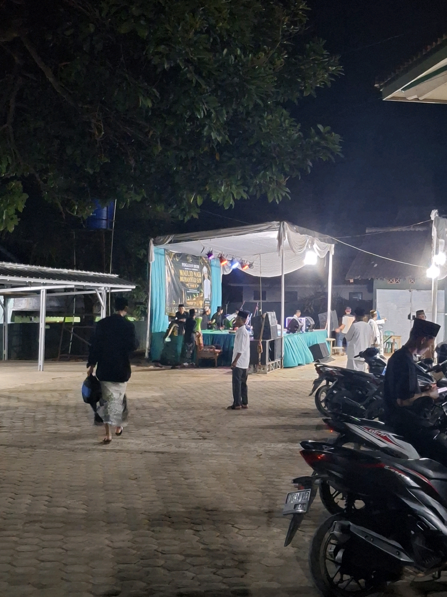 This is a medium shot, low-angle photograph taken at night in what appears to be an outdoor event space in Yogyakarta, Indonesia. The scene is illuminated by artificial lighting from stage lights and overhead lamps. In the foreground, several parked motorcycles are visible. A person in the middle ground, wearing a black jacket and a sarong, is walking away from the camera, carrying a blue bag. To the right, a person in a white shirt and dark pants is standing. In the background, a stage is set up with a white canopy, decorated with a teal banner displaying text in Indonesian, likely related to a religious event. Several people are gathered on and around the stage, some appearing to be performers or speakers. The ground is paved with interlocking bricks. A large tree with dense foliage is visible in the upper left quadrant, casting shadows. The overall atmosphere suggests a community gathering or religious celebration taking place in an urban or semi-urban setting.
