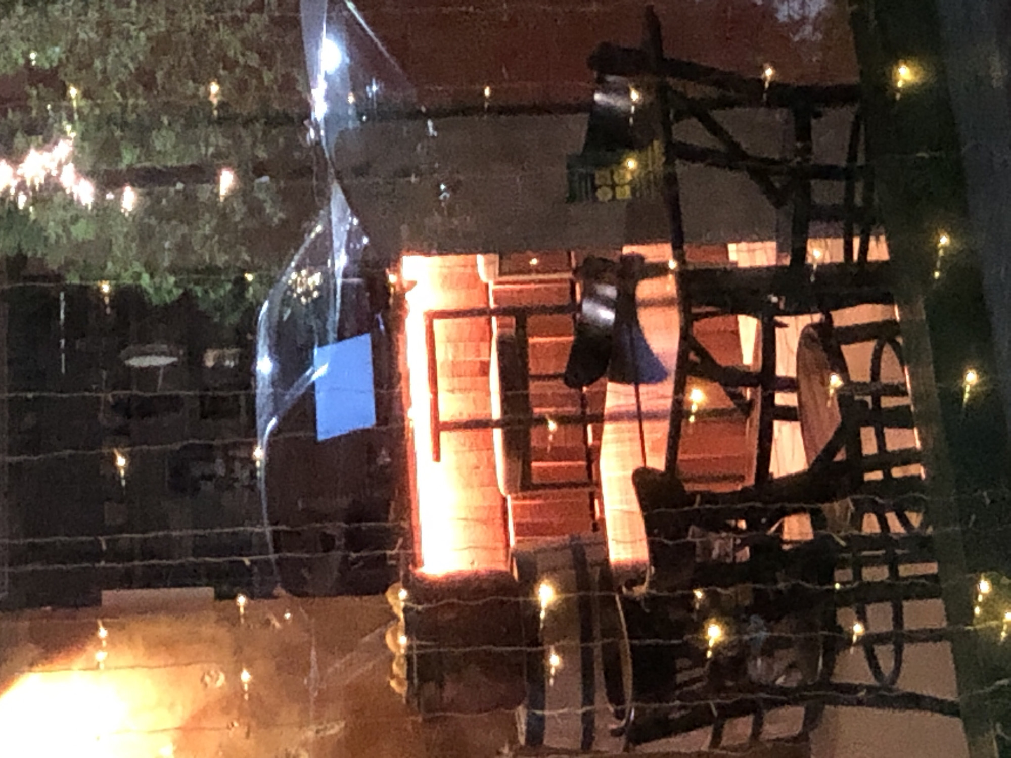 The image captures a dimly lit outdoor seating area, possibly a patio or a section of a restaurant, at night in Saint Petersburg, Russia. String lights, some with a warm yellow glow, are draped across the scene, creating a festive yet somewhat hazy atmosphere.
In the foreground, a dark wooden table with a rustic design is visible. It appears to be set for patrons, with several chairs around it. The chairs are also made of dark wood, with intricate detailing in their construction, suggesting a stylistic choice for the establishment. Some chairs are empty, while others appear occupied by dark, indistinct shapes, possibly people or bags.
Behind the table, stacked wooden barrels add to the rustic ambiance. The barrels appear to be used as decorative elements or possibly to hold items. A warm, orange light emanates from behind these barrels, illuminating them and casting a reddish hue on the surrounding area.
Further in the background, the sleek, dark silhouette of a car is visible, partially obscured by the overall darkness and reflections on a window. The car has a blue illuminated element on its dashboard or windshield, catching the light. The presence of the car suggests the scene is located near a street or a parking area. Trees with dense foliage, also illuminated by some light sources, are seen beyond the car, indicating an outdoor setting within a city.
The overall impression is one of a cozy, perhaps slightly dimly lit, outdoor dining or drinking establishment. The reflective surfaces, possibly a window or a glass enclosure, create a layered effect, showing both the interior of the establishment and elements from the exterior. The lighting, especially the string lights and the warm glow from behind the barrels, suggests an evening setting intended to create a welcoming and intimate mood. The environment appears to be an urban setting within Saint Petersburg, characterized by the presence of cars and greenery.