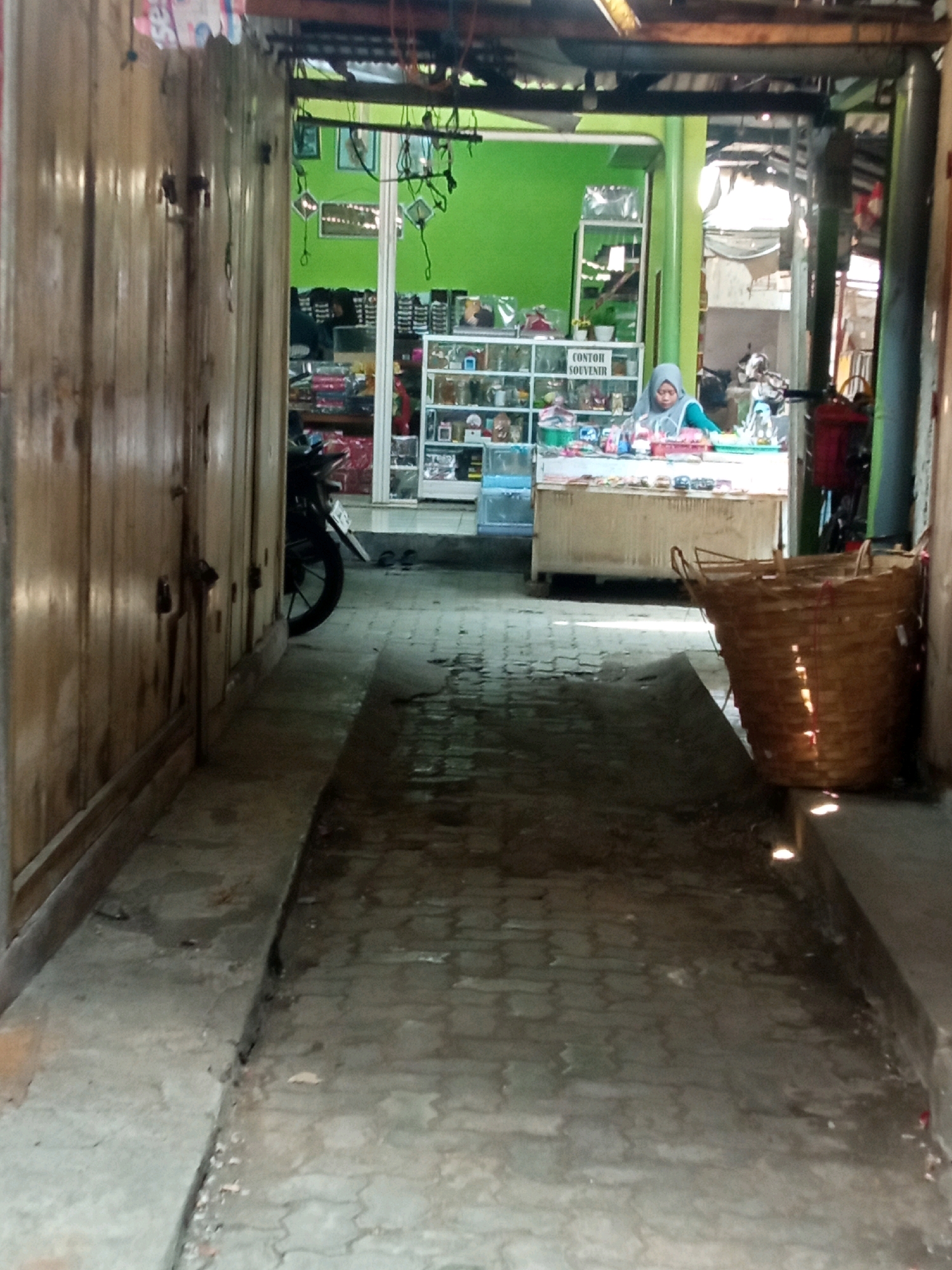 The image depicts a narrow, covered walkway or alleyway in Sambong, Indonesia, leading to an open-air shop. The walkway is paved with interlocking bricks and is flanked by wooden walls on the left and a concrete structure on the right. A motorcycle is parked on the left side of the walkway, partially obscured by the wooden wall.
In the background, a shop with bright green walls displays various items on shelves and a counter. A woman wearing a hijab is standing behind the counter, seemingly attending to customers or arranging merchandise. Various goods, including what appear to be souvenirs and packaged items, are visible on the shelves and counter. A large woven basket is placed on the right side of the walkway, in front of the shop. The lighting suggests an outdoor or semi-outdoor setting, with sunlight filtering through the covered structure and reflecting on the wet pavement.
