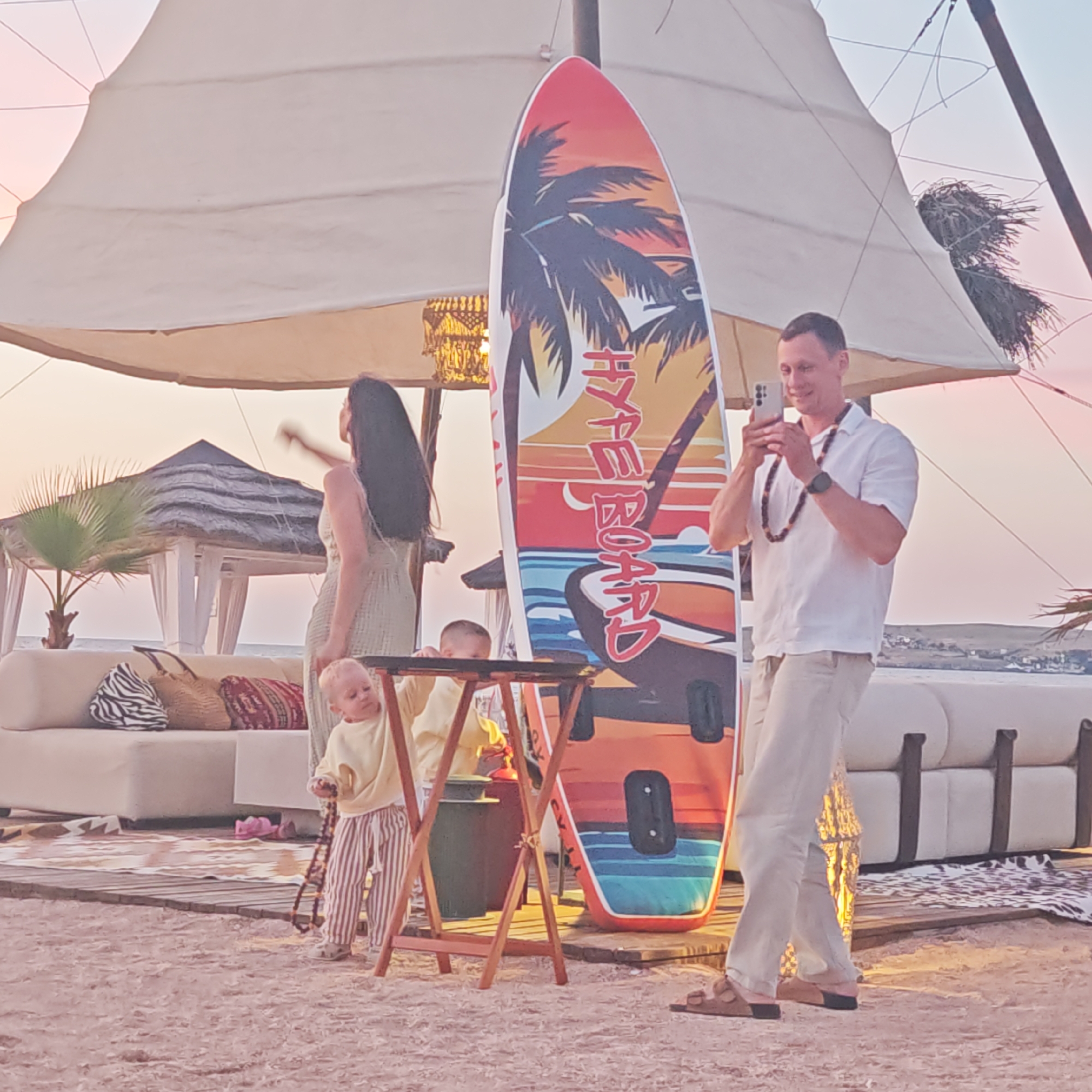 In Мысовское сельское поселение, Ukraine, at dusk, a man in a white shirt and tan pants stands on a sandy beach, holding a smartphone to take a picture. He wears a dark, beaded necklace. To his left, a woman with long dark hair, wearing a light-colored dress, stands with her arm raised. In front of them, a young child in a yellow sweatshirt and striped pants holds a beaded necklace. A surfboard with a tropical sunset design stands upright nearby. The scene is set under a large, white canopy, with lounge furniture and pillows visible in the background. A small, thatched-roof structure and palm trees are also present. The sky exhibits soft pink and orange hues characteristic of sunset.
