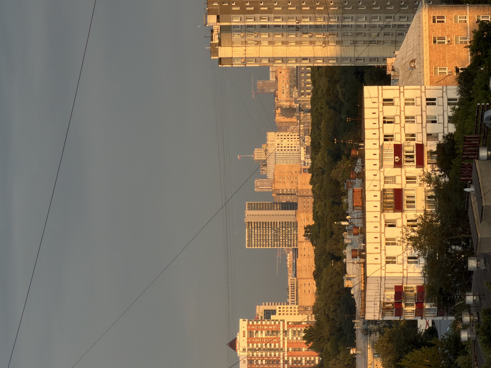The image captures a cityscape during sunset in Khimki, Russia. The golden hour light illuminates several residential buildings of varying heights and architectural styles. In the foreground, a white building with red balconies is lit from the side, showcasing its repetitive window patterns. To the right, a taller, grey residential building with a similar window structure is also bathed in the warm light. In the background, a cluster of buildings, including a distinctive orange and white high-rise with a pyramidal roof, stand against a grey, overcast sky. A line of dense green trees runs horizontally across the mid-ground, separating the lower buildings from the taller ones. Overhead, power lines stretch across the frame, adding a sense of urban infrastructure. The overall impression is one of a typical urban residential area experiencing the end of a day. There are no visible people or specific actions depicted.