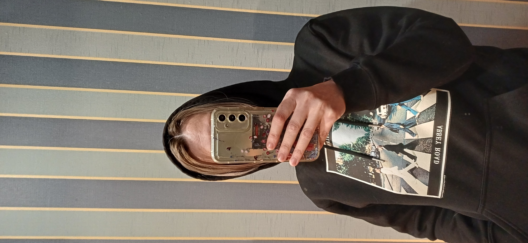 A person is taking a selfie with a phone in a decorative case. The person is wearing a black hooded sweatshirt with an "Abbey Road" album cover design on the front. The background consists of horizontal stripes in shades of blue, gray, and gold. The location is not specified or determinable from the image.