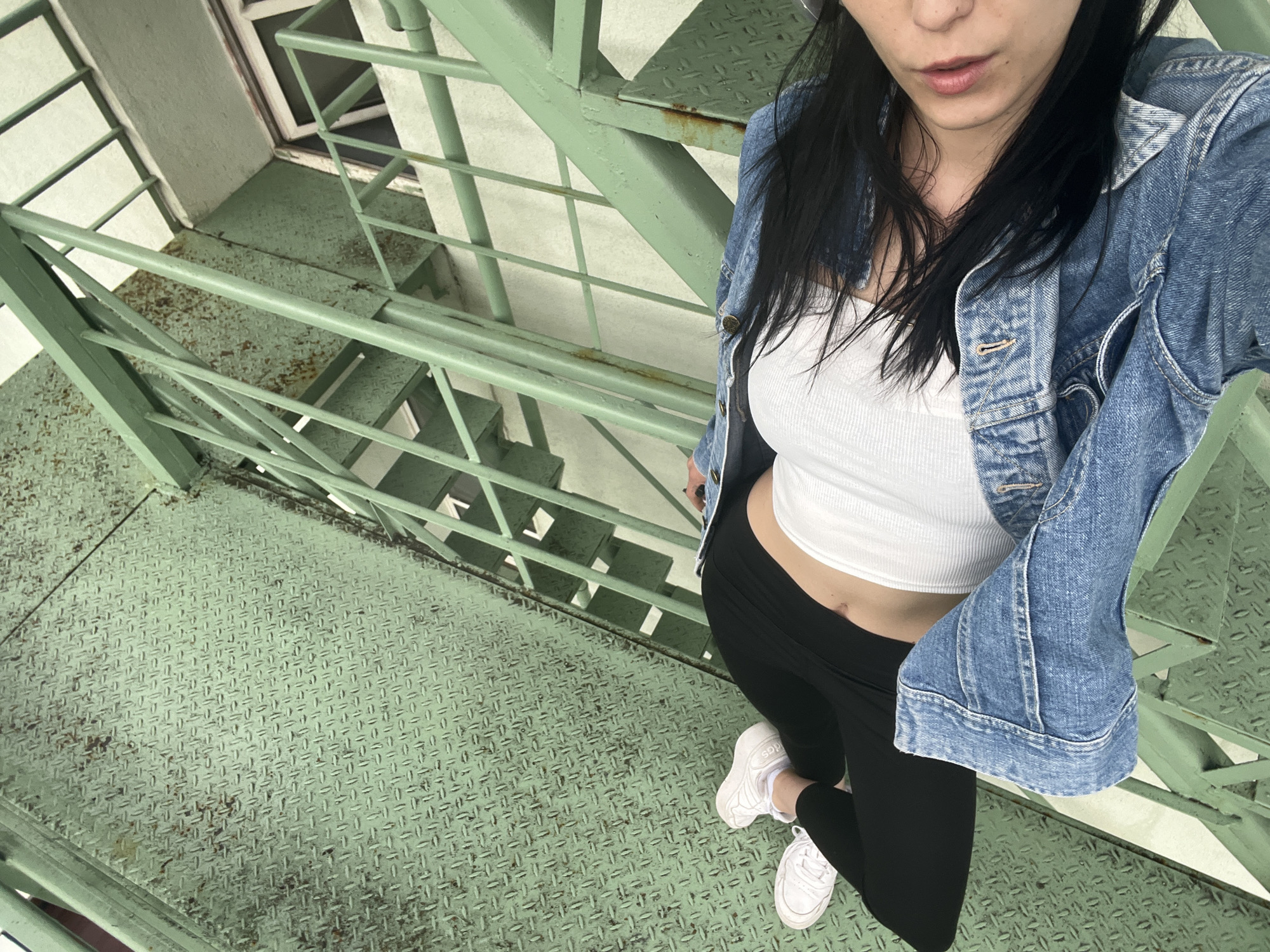 A medium-angle, overhead shot captures a woman standing on a worn, green metal staircase. The woman is partially visible from her lower face (mouth and nose) down to her upper thighs and feet. She has long, dark hair and is dressed in a light blue denim jacket over a white ribbed crop top that exposes her midriff, black leggings, and white sneakers. The word "das" is visible on the side of one of her sneakers, likely indicating the brand Adidas.
The staircase, possibly an outdoor fire escape, is made of diamond plate metal treads and railings, all painted a faded green with noticeable rust and scuff marks in various areas. The perspective looks down the stairs, revealing several flights below. In the background, parts of a light-colored building wall and window frames are visible. The lighting suggests it is daytime. The woman's expression is neutral.