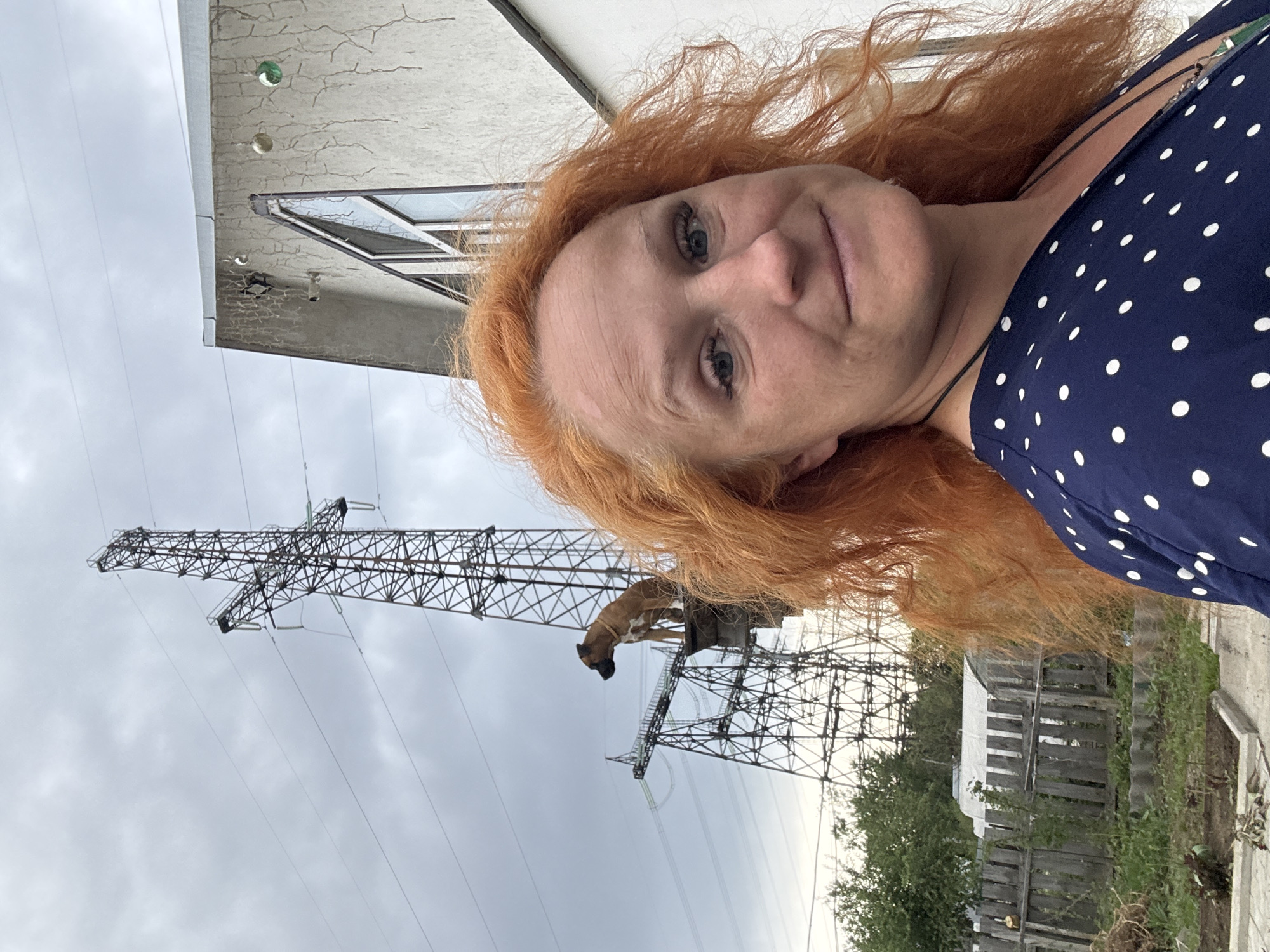 The image depicts a woman with red hair wearing a blue and white polka dot dress, presumably taking a selfie. In the background, there's an electrical transmission tower with a dog standing atop its base, likely in a residential area. The sky is overcast. The location is indicated as Chaikovsky, Russia. There is a house on the left with a window and some landscaping with trees and an old wooden fence on the right.
