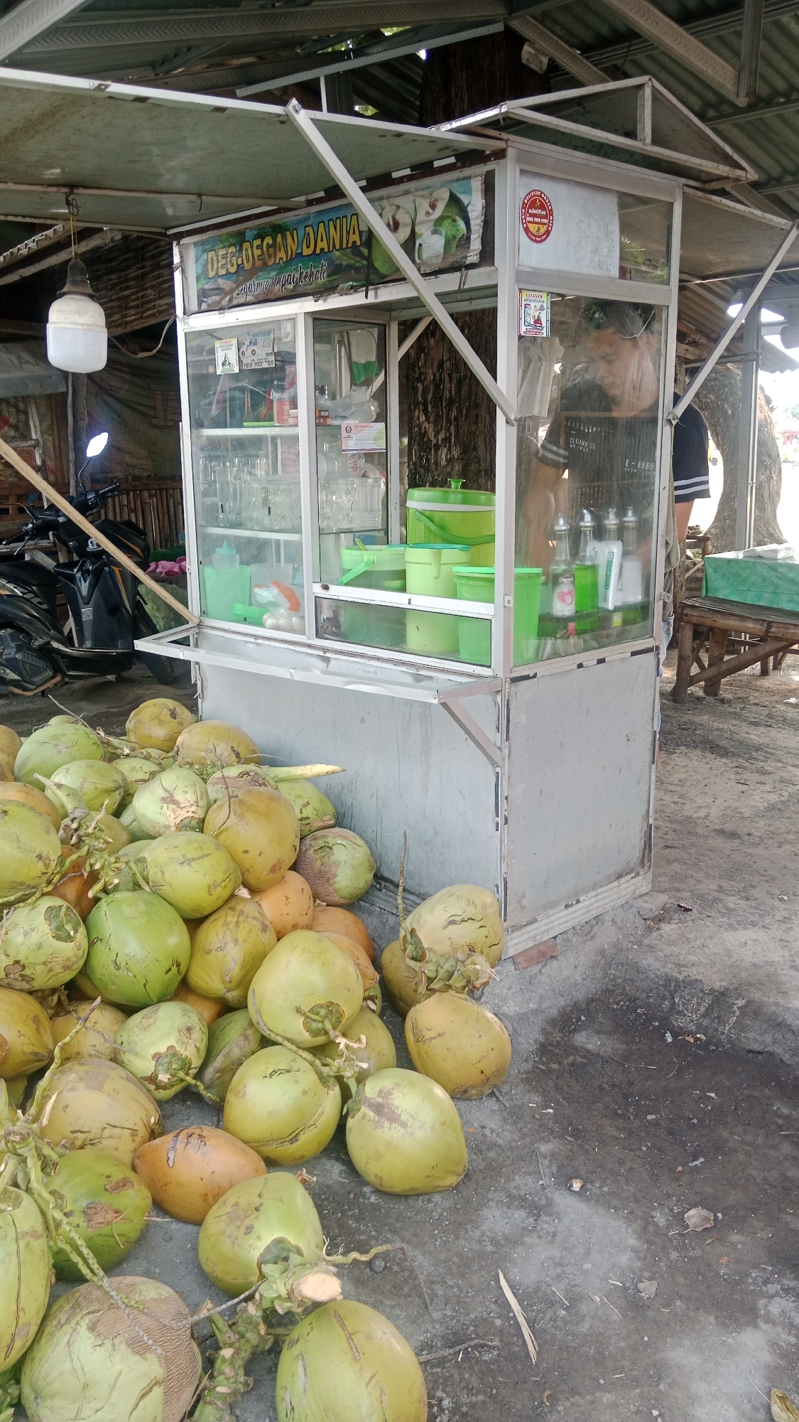 In Patunjungan, Indonesia, a street vendor stands behind a glass-enclosed beverage cart, serving drinks. The cart is positioned on a sandy ground, under a simple, open-air roof supported by metal poles. A pile of green and brown coconuts sits in front of the cart, suggesting the nature of the business. The cart itself is gray and white, with a sign above the glass display that reads "DEG-DEGAN DANIA". Inside the cart, various bottles of syrup and cups are visible, along with green buckets. A motorcycle is parked to the left of the cart, partially obscured by a bamboo-like structure. In the background, a large tree trunk is prominent, and a wooden bench with a green tarp sits to the right. The scene appears to be outdoors, possibly in a market or along a roadside, with natural lighting.