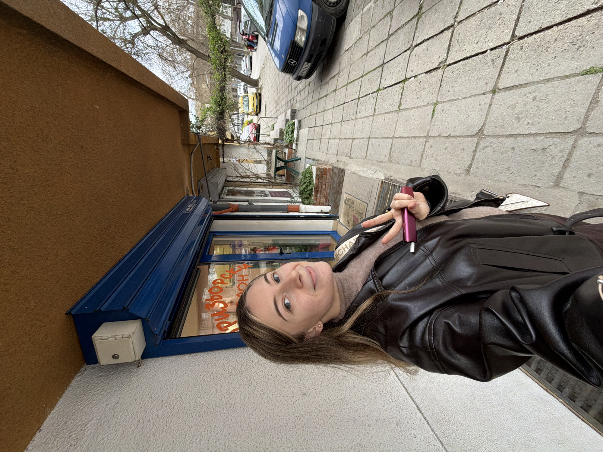 A young woman stands on a sidewalk next to a building with a blue awning and a sign in Bulgarian. She is wearing a brown leather jacket and holding a pink object in her hand. The sidewalk is paved with gray stones. A blue Mercedes is parked on the side of the road. Trees and other buildings are visible in the background. The location is Plovdiv, Bulgaria.