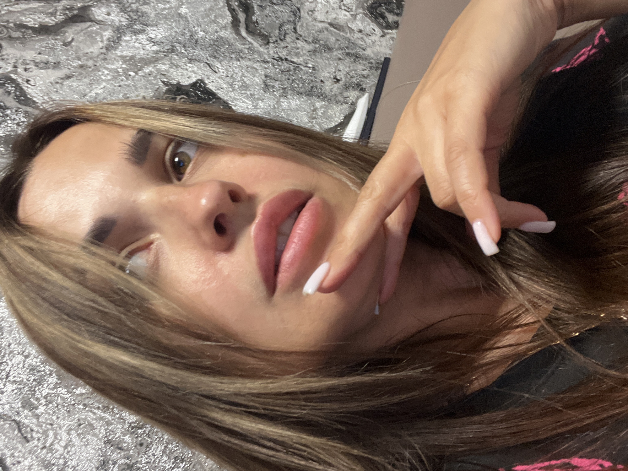A woman with shoulder-length, light brown hair and prominent eyebrows is captured in a close-up, overhead shot. Her left hand, with neatly manicured light pink nails, is raised to her chin, with her index finger resting on her lower lip. Her mouth is slightly open, revealing her teeth, and her eyes are looking to her left. The background is a textured, metallic silver wall, and a beige surface is visible in the lower right corner. The woman is wearing a black garment with a pink pattern. The lighting suggests an indoor setting, possibly a home or studio.