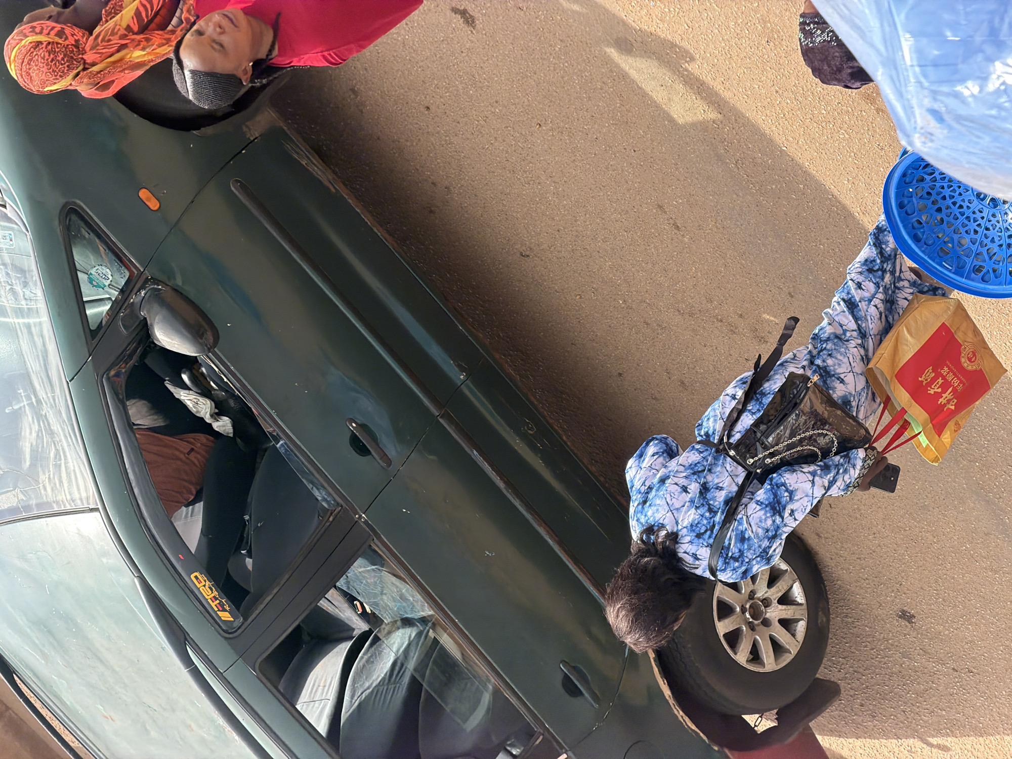 In Bukuru, Nigeria, a dark green four-door sedan is parked on a dirt road. Two women are visible. One woman, wearing a bright red shirt and a patterned headscarf, sits in the passenger seat of the car. Another woman, wearing a blue and white tie-dye outfit, stands on the ground near the car's rear wheel. She is holding a cell phone and two shopping bags, one red with Chinese characters and one yellow. A blue plastic basket is also on the ground near her. The car has a dent on its side and some damage to the windshield. The overall lighting suggests it is daytime.