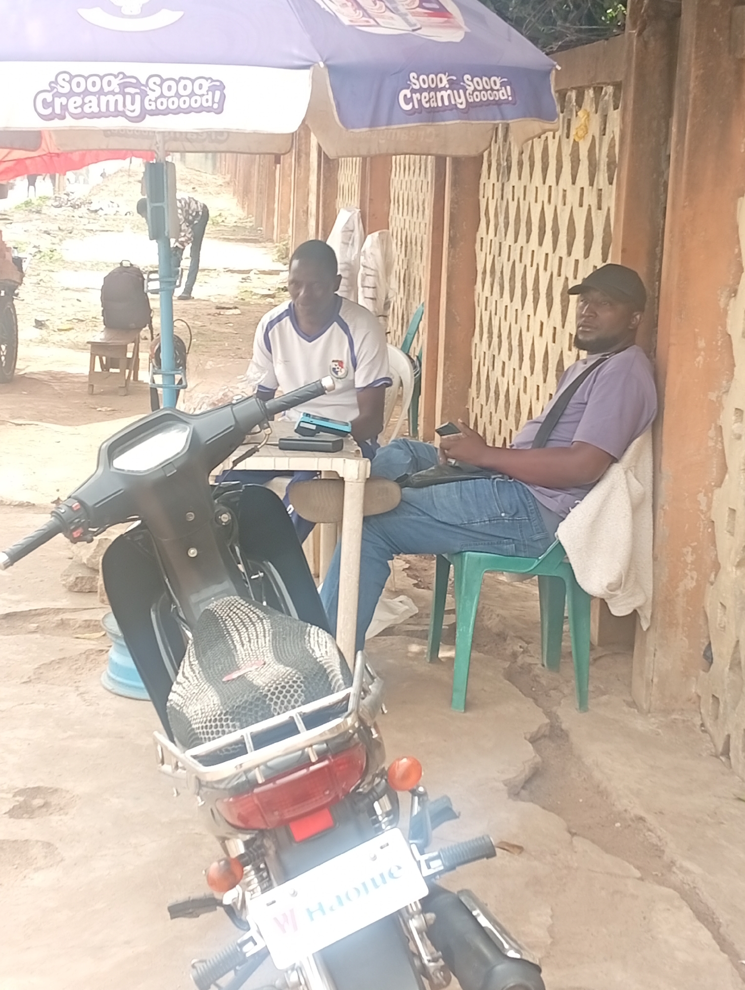 This outdoor image, taken in Jos, Nigeria, depicts two men relaxing under a large umbrella. In the foreground, a black motorcycle is parked, partially obscuring the man on the left. The motorcycle features a mesh seat cover and a rear rack; its license plate is partially visible, showing "W Haoue".
To the left, a man in a white and blue shirt sits at a small white table, appearing to be using a mobile phone. Behind him, a black backpack rests on a wooden stool. To the right, another man, wearing a purple shirt, blue jeans, and a dark cap, is seated on a green plastic chair, leaning against a cream-colored, patterned wall. A white cloth is draped over the back of his chair, and he is also holding a mobile phone. Both men appear relaxed.
Above them, a large umbrella provides shade, displaying the text "Sooo Creamy Goooood!" The ground is unpaved, appearing dusty or sandy. In the distant background, another individual can be seen bending over. The scene suggests a casual, street-side setting during daytime, possibly a break from activities or a common gathering spot.