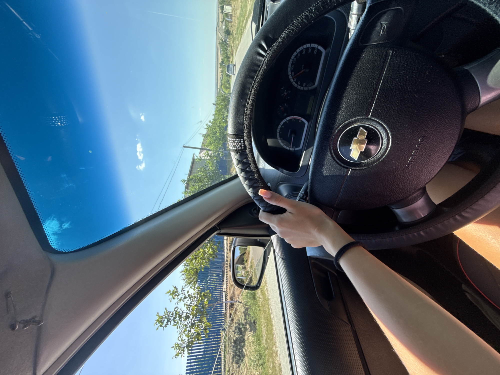The image shows the interior of a car, with a driver's hand on the steering wheel. The driver is holding the steering wheel with their right hand, and their left arm is resting on the car door. The steering wheel has a black cover with rhinestones, and a Chevrolet logo in the center. The dashboard shows a speedometer and a tachometer. Outside the car, there is a road with a few houses and trees. The sky is blue with some clouds. The scene is likely in a rural area near Basarabeasca, Moldova, given the style of houses and fences. The lighting suggests it is daytime, likely late morning or early afternoon.