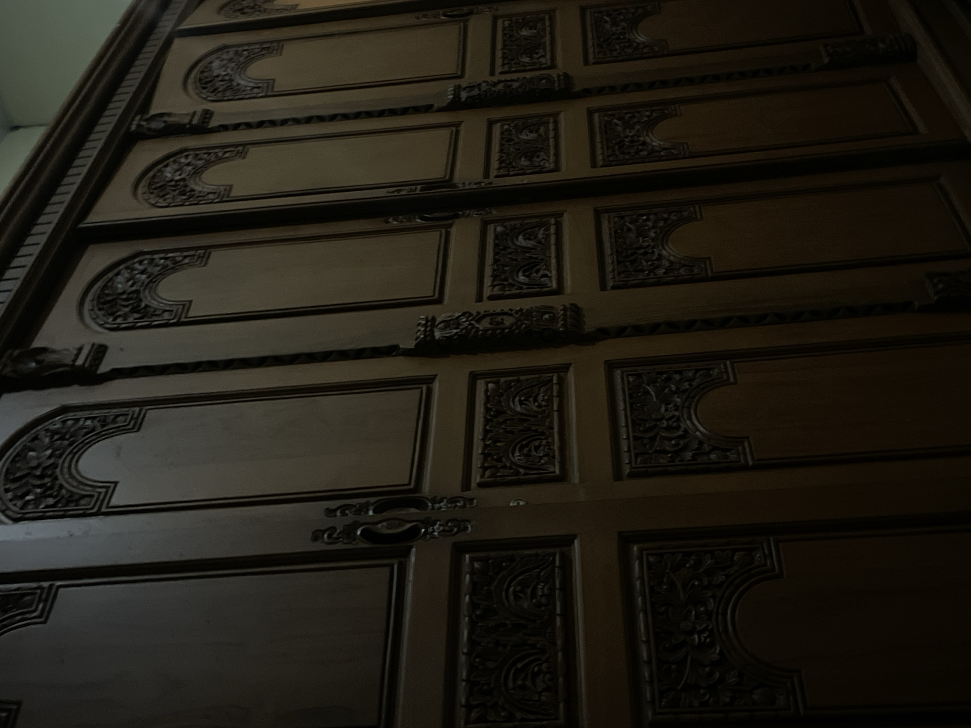 The image shows a close-up of a dark wooden wardrobe with intricate carvings on its doors. The wardrobe appears to be large, with multiple doors visible in the frame. Each door has a distinct design, featuring arched panels with detailed floral and geometric patterns carved into the wood. The handles are also ornate, made of dark metal. The lighting in the image is dim, casting shadows that highlight the texture and depth of the carvings. The overall impression is of a traditional and well-crafted piece of furniture. The location in Manado, Indonesia, is not evident from the image itself.