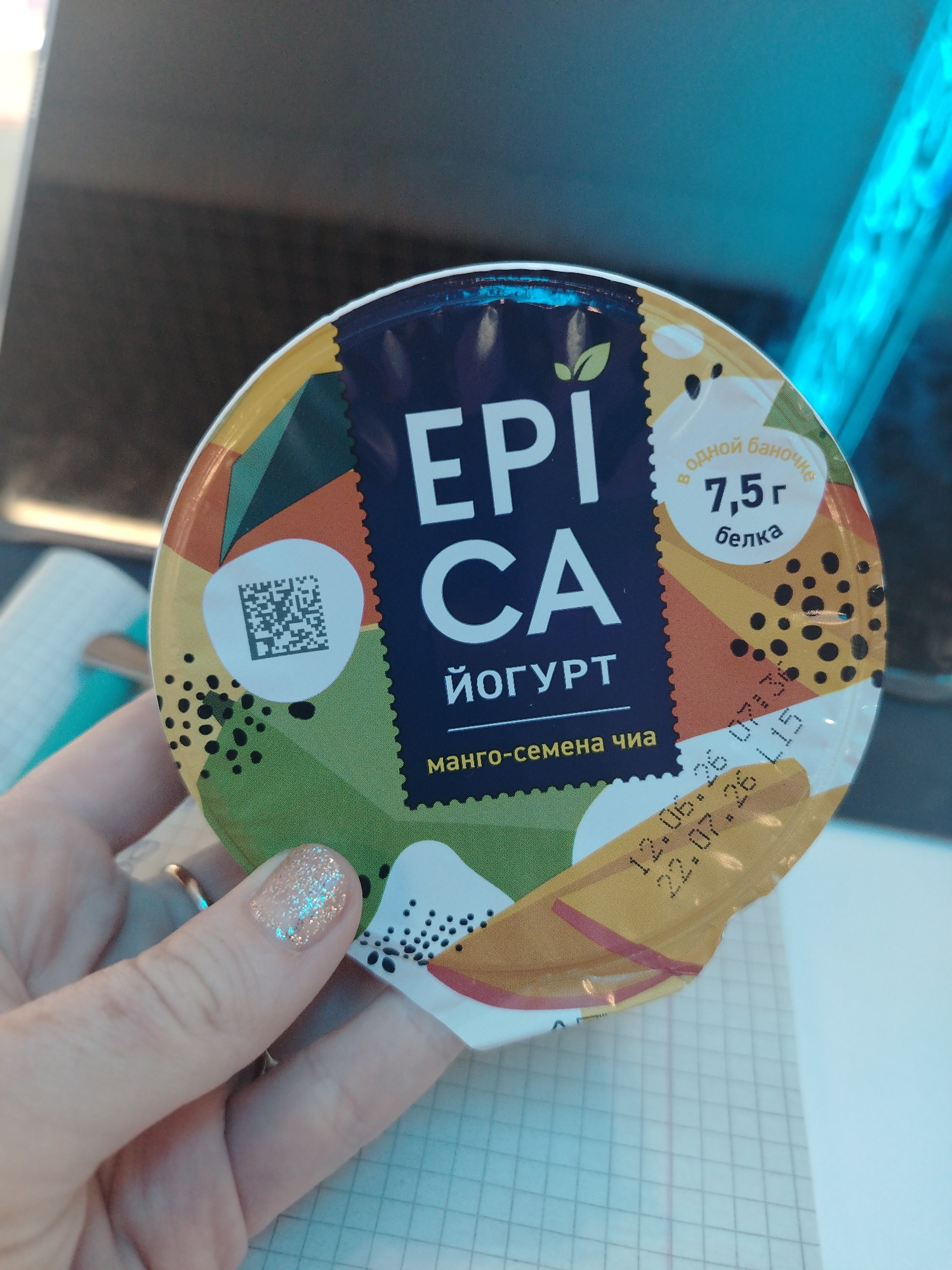 The image shows a hand holding a round yogurt container with colorful branding. The text on the container is in Russian and includes the brand name "EPICA" and "ЙОГУРТ." There are also details about the flavor, "манго-семена чиа" (mango-chia seeds), and nutritional information, "7,5 г белка" (7.5g protein). A QR code is visible on the container. The background is blurred but appears to be an indoor setting with a dark surface and a bright blue, possibly illuminated object. There are no discernible landmarks or details to suggest a specific location in Izhevsk, Russia.