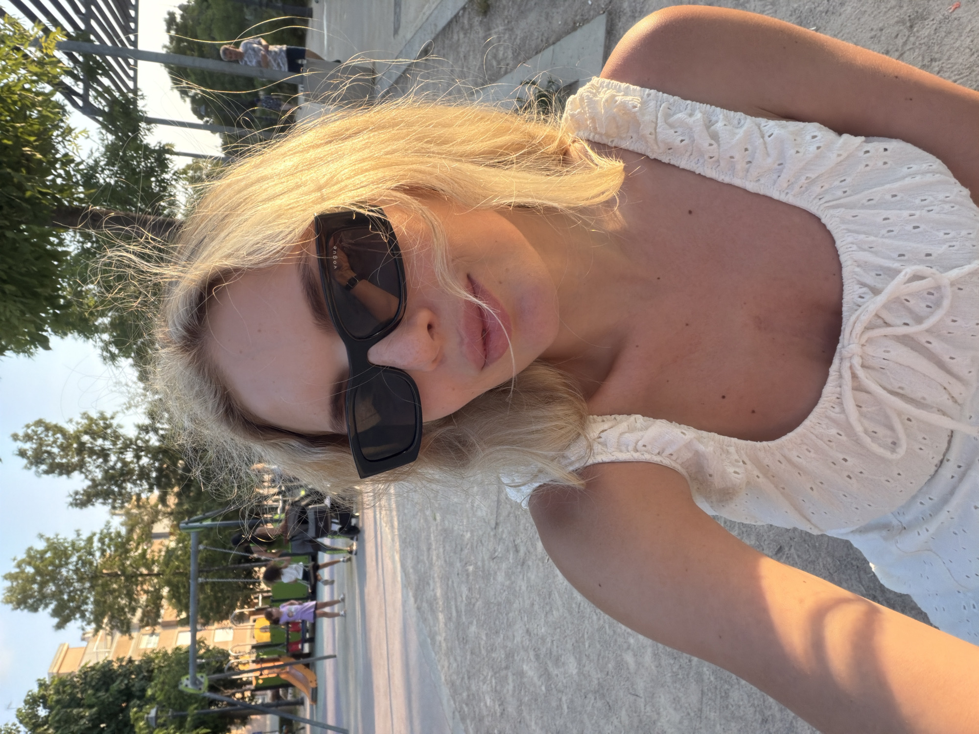 The image shows a close-up selfie of a young woman with blonde hair and sunglasses, standing in an outdoor public space. She is wearing a white, eyelet-fabric top with ruffled straps and a drawstring tie at the bust. The background reveals a playground with swings and slides, children playing, and trees. In the distance, there are buildings and a pedestrian walkway with modern, slatted overhead structures. The lighting suggests it is late afternoon or early evening, with warm sunlight casting shadows. The setting appears to be a park or recreational area within a residential urban environment. Based on the provided information, the location is identified as Benimàmet-beniferri, Spain.