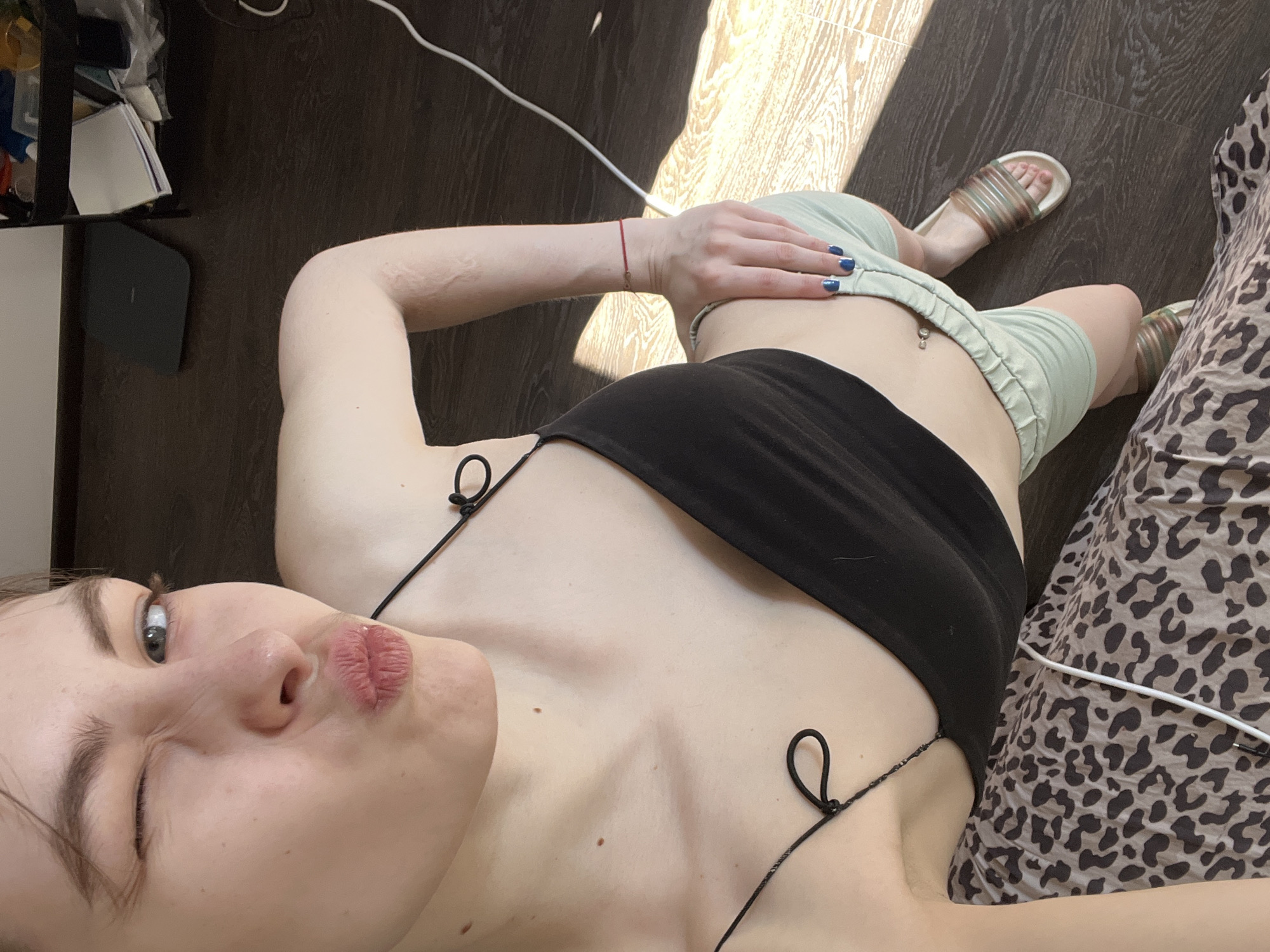A woman lies on her side, facing the camera, with her left eye slightly closed and her lips pursed. She wears a black top with thin straps. She is wearing green shorts. Her hand rests on her abdomen, with her fingers displaying blue nail polish. A red string is visible on her left wrist. She is wearing sandals. The background shows a wood-patterned floor, a section of a bed with animal print bedding, and a white cable. A portion of a table and its contents are visible in the top left corner. There is nothing to indicate that the image was taken in Saint Petersburg, Russia.