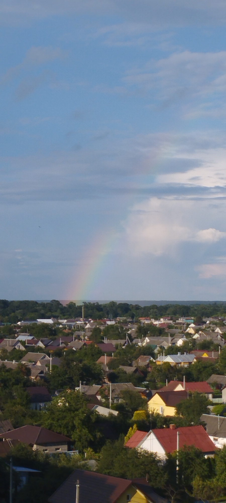 An aerial perspective captures a residential area in Skidzieĺ, Belarus, under a partly cloudy sky with a rainbow arching across the horizon. The scene is dominated by a dense cluster of houses with varied roof colors, including grey, brown, red, and yellow, nestled amidst lush green trees. Roads and power lines are visible throughout the neighborhood. In the distance, a treeline separates the settlement from the sky. The image appears to be taken during daylight hours, possibly late afternoon or early evening, judging by the lighting. There are no people visible in the frame.