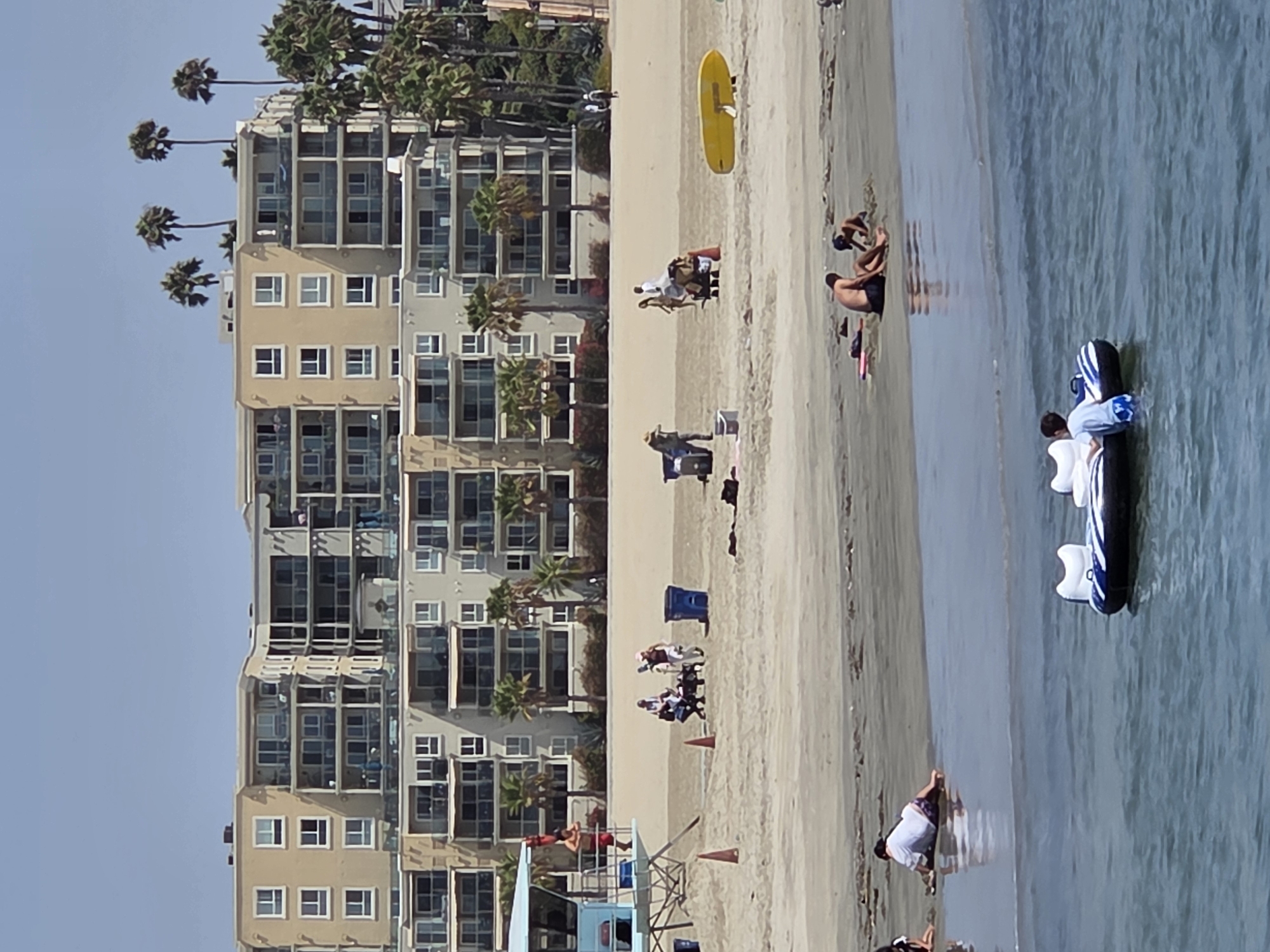 The image captures a lively scene at a beach in Long Beach, California. In the background, a multi-story apartment building with balconies and numerous windows stands prominently, partially shaded by palm trees. The beach itself is populated with various individuals engaged in recreational activities.
In the foreground, a blue and white inflatable raft floats in the shallow water, with one person standing beside it. Further back, closer to the shore, a group of people are spread out on the sand. Some are lounging, while others appear to be building sandcastles. A yellow surfboard is propped up on the sand.
A lifeguard tower, painted light blue, is positioned on the beach, with a lone figure standing on its platform. In the mid-ground, several people are interacting, some pushing a cart laden with belongings. Trash cans are visible on the sand, suggesting a maintained public space. The overall impression is one of a sunny day at a popular beach destination.