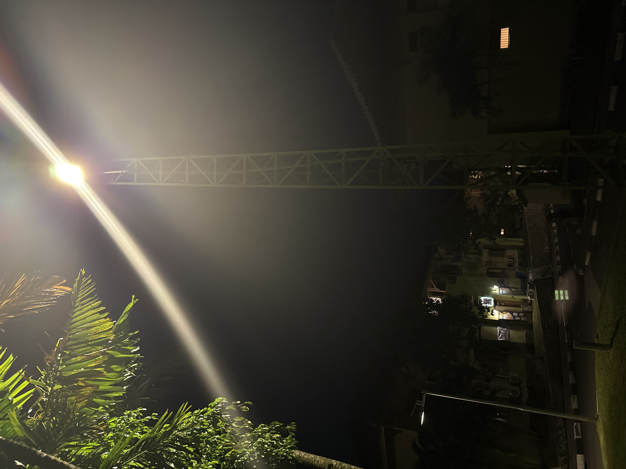 This media file captures an outdoor night scene, likely in an urban or suburban setting, under artificial illumination. The dominant subject is a very bright street light positioned on the left side of the frame, casting a strong, elongated light flare diagonally across the upper left and central portions of the image.
Below and to the left of the light source, lush green tropical foliage, including large palm fronds and other dense plants, is heavily illuminated. Extending horizontally across the middle of the frame is a long, light-colored metal truss structure, possibly part of a pedestrian bridge, an overhead walkway, or a utility support.
To the right, a multi-story building with light-colored (possibly light green or beige) walls is visible. Several windows on the building are illuminated from within, and multiple external air conditioning units are mounted on its facade. Below the building, there's a paved path or road bordered by distinctive black and white striped curbs, alongside a neatly maintained grassy area. Another, less prominent street light pole can be seen in the lower right corner.
The sky is dark, indicating nighttime, with some atmospheric diffusion around the powerful street light. No people are visible, and no discernible activities are taking place. The overall impression is a quiet, artificially lit nightscape. While there are signs and banners on the building in one of the crops, no text is clearly legible. The presence of tropical foliage aligns with the location context of Kuta Selatan, Indonesia.