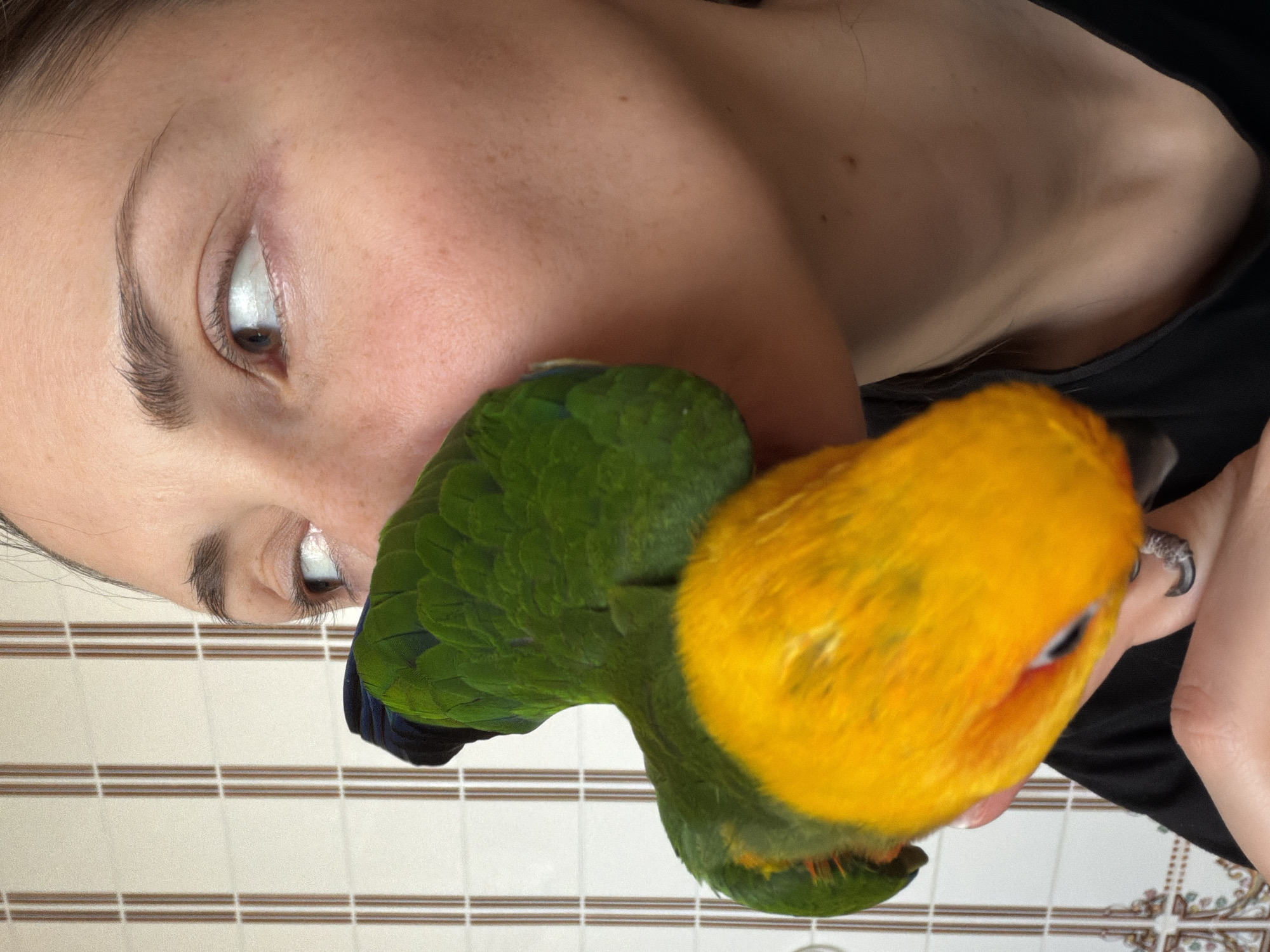 A close-up, vertical photograph captures an intimate interaction between an adult female and a parrot within an indoor setting, reportedly in Khimki, Russia. The female, visible from the mid-chest upwards, presents her right profile, with her head slightly turned to the right. She has light skin exhibiting visible freckles, and her right eye, featuring a brown iris, is open and directed towards the right. Her hair appears dark and is pulled back or short. She wears a dark, possibly black, garment. A parrot, characterized by green plumage on its back and wings, and bright yellow-orange coloration on its chest and face, is positioned against the female's right cheek. Its head is largely obscured by the contact point with her cheek. One of the parrot's feet is visible gripping the female's right hand, which is supporting the bird from below. The background consists of an off-white tiled wall featuring vertical brown striped patterns. Decorative tiles with a floral or ornamental motif in brown and beige tones are visible on the lower portion of the wall.