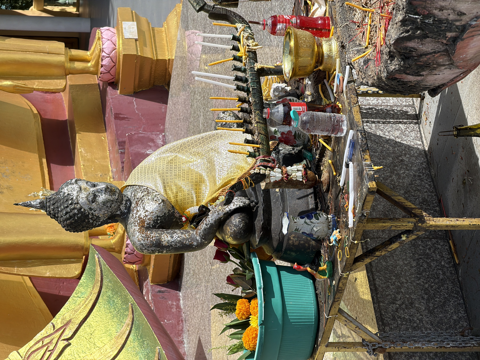 In Baan Plai Laem, Thailand, a weathered stone Buddha statue, draped in a golden cloth, sits in front of a larger golden statue. The scene is set on a paved area, likely within a temple complex, given the presence of multiple religious offerings. A table to the left of the Buddha holds a teal plastic basin filled with orange marigolds and red roses, a small colorful rooster figurine, and a decorative vase. In front of the Buddha, a metal stand displays numerous lit and unlit yellow and white candles, some with melted wax dripping down. A golden bowl, a red bottle, a bottle of water, and a bottle of dark soda are also on the table. To the right, a large stone incense burner is filled with lit red incense sticks. The background features a large golden structure with intricate detailing and decorative bases.