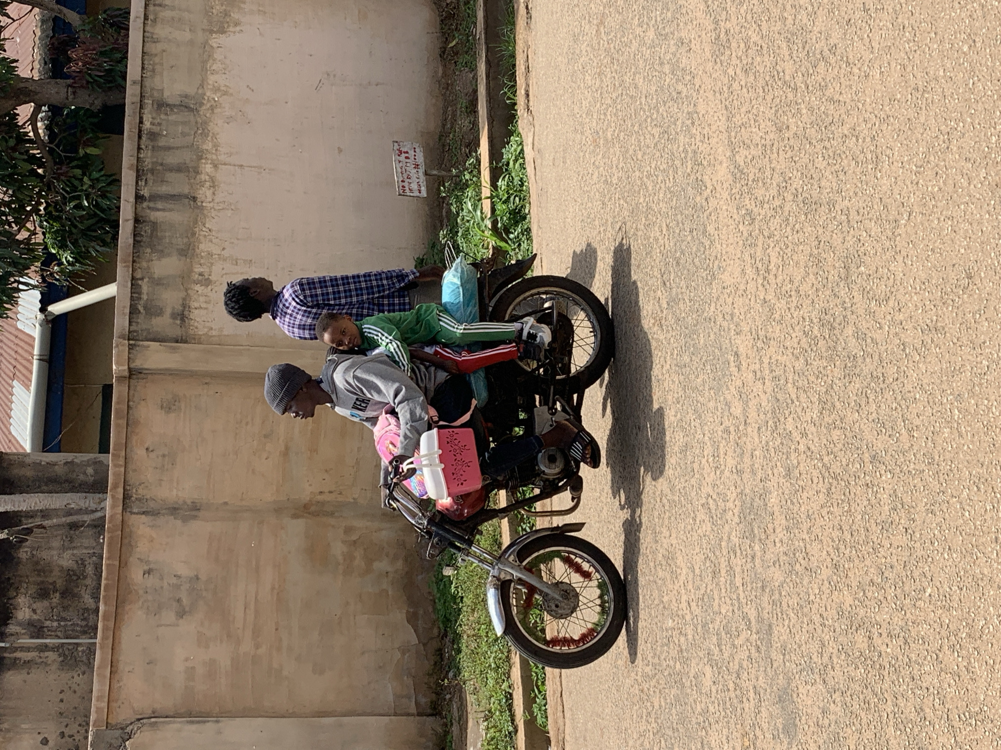 The image shows three individuals and a motorcycle on a sunny day in Jos, Nigeria. An adult male in a purple and white plaid shirt stands behind a black motorcycle. Seated on the motorcycle, positioned in front of the driver, is a child wearing a green tracksuit with white stripes. The driver, an adult male in a grey hoodie and a dark beanie, is seated on the motorcycle, holding the handlebars.
The motorcycle has a red fuel tank, and a pink container along with a white object are secured near the handlebars. The wheels of the motorcycle have red decorative elements. The individuals appear to be stationary, possibly preparing to depart or having just arrived.
The background consists of a tall, light-colored wall showing signs of weathering and dark stains. Green vegetation, including a small tree, grows along the base of the wall. On the far left, a tiled roof and a white gutter are partially visible. The ground is a light-colored, textured surface, likely a paved road or path.
A small, hand-painted sign is visible amidst the vegetation. Partially legible text on the sign includes "No...", "DJ MOB", and "House" along with some numbers. The strong sunlight creates clear shadows, suggesting it is daytime, likely morning or afternoon.