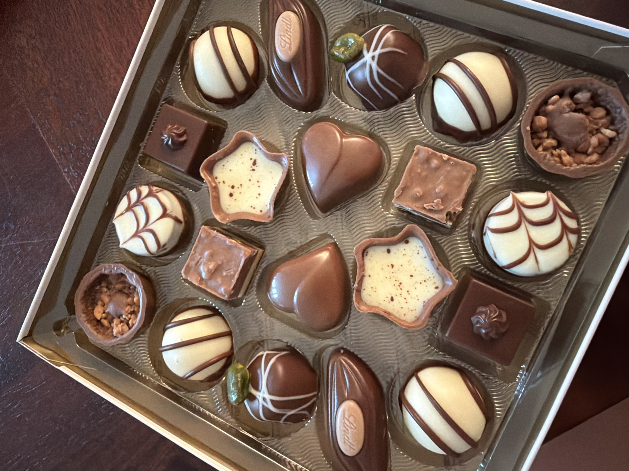 The image is a top-down, close-up view of an open box of assorted Lindt chocolates. The chocolates are arranged in a grid within a clear plastic divider. The chocolates vary in shape and decoration, including white chocolate with dark chocolate stripes, dark chocolate squares with molded tops, heart-shaped chocolates, star-shaped chocolates with speckled filling, a chocolate shell filled with nuts and caramel, and a torpedo-shaped chocolate with a Lindt logo. The box itself has a gold-colored border. The background appears to be a dark wooden surface. The image was taken in the city of Martigny, Switzerland, likely in a domestic setting such as a home or office, as indicated by the presence of food items in a box. There are no people or actions depicted in the image.