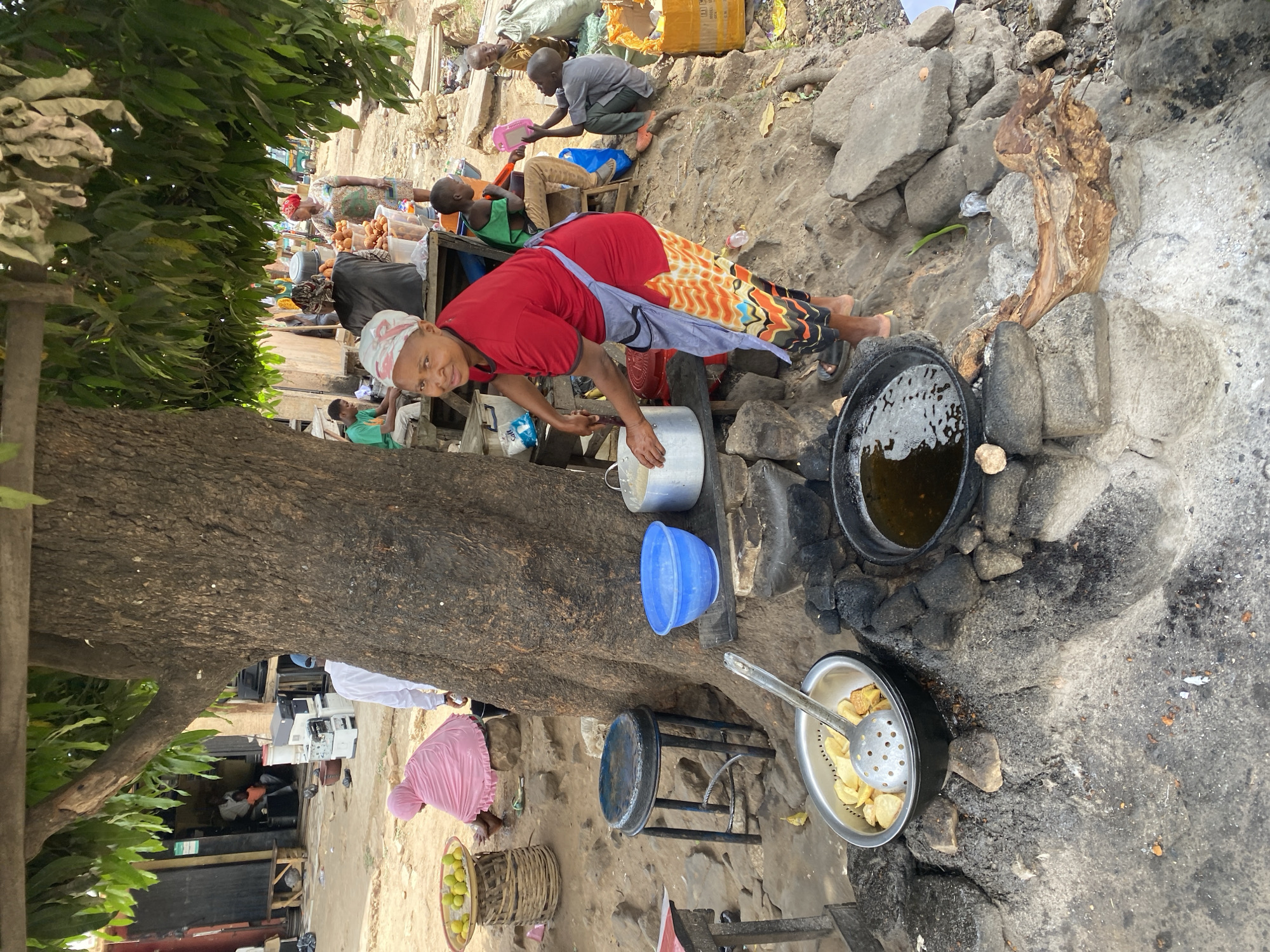 The image captures a street food vendor in Jos, Nigeria. A woman wearing a red shirt, patterned skirt, headscarf, and apron is shown preparing food. She is holding a silver pot, positioned near a large tree trunk. A black pot of oil sits atop a makeshift stone stove, next to a bowl of fried food with a slotted spoon. In the background, several people are seated and engaged in conversation or activities, while others are standing nearby. The environment appears to be an outdoor market or commercial area.