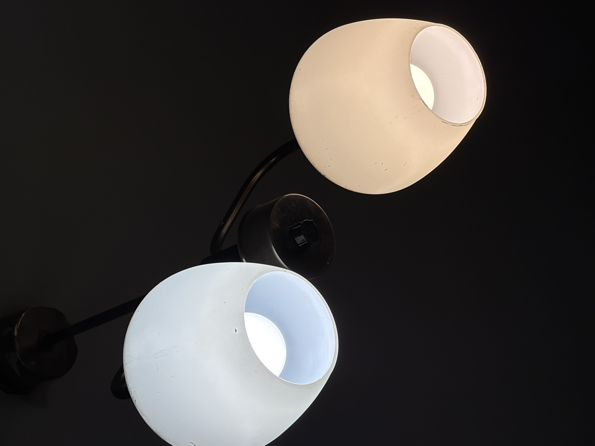 A close-up, low-angle shot captures two illuminated lampshades against a dark background. The lamp on the left emits a bright white light, while the one on the right glows with a warmer, yellowish hue. Both shades are opaque and have a rounded, somewhat elongated shape. A dark, metallic fixture connects the lampshades to what appears to be a ceiling. The image is devoid of any contextual clues that would indicate a specific location within the city of Sofia, Bulgaria, or any identifiable individuals or actions.