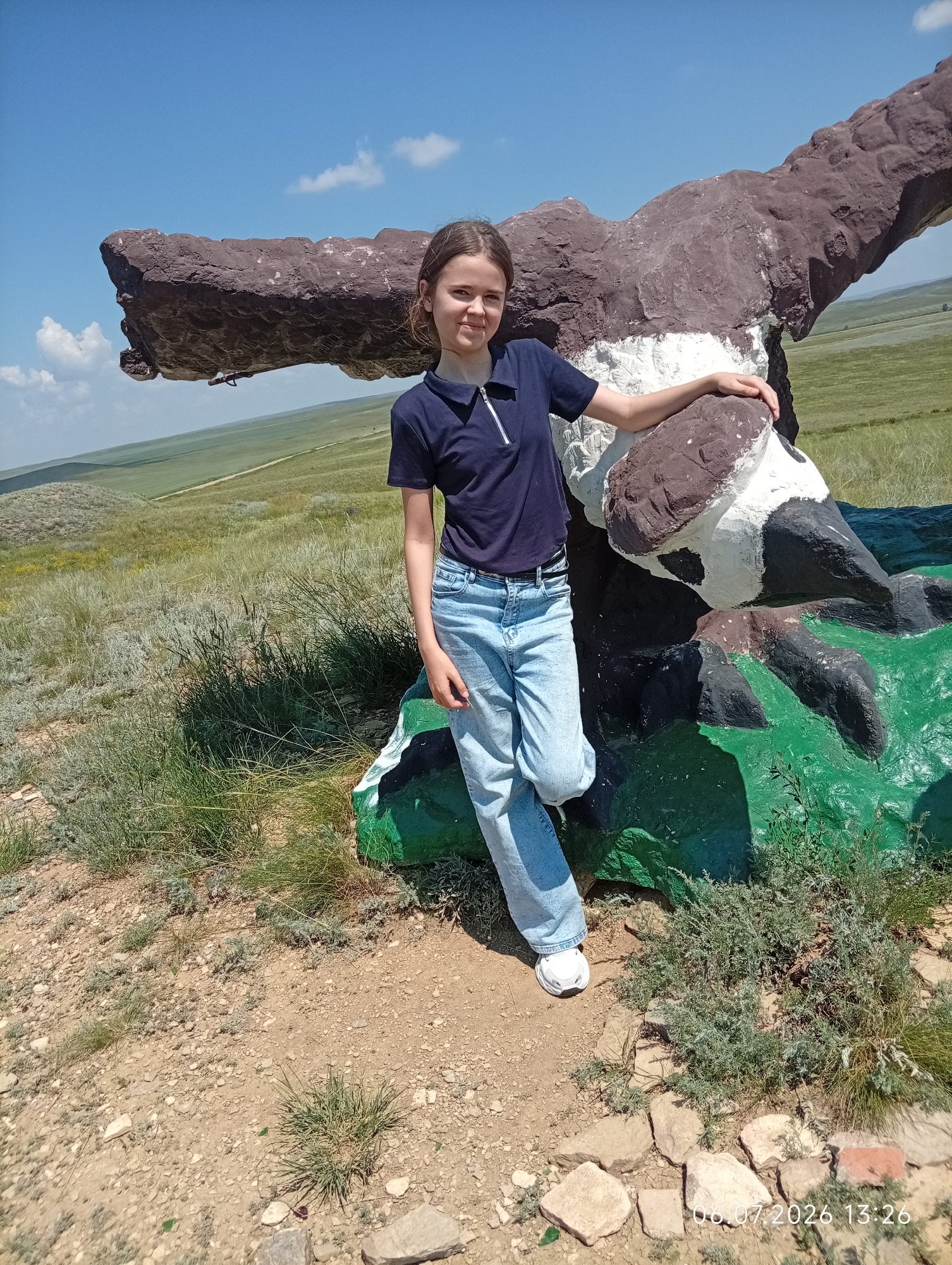 In Aleksandrovka, Kazakhstan, a young girl stands in front of a large, sculpted rock formation resembling an eagle in flight. The girl, with brown hair and wearing a dark blue polo shirt and light blue jeans, is leaning against the eagle's wing. The rock sculpture is painted in various earthy tones of brown, white, and black, with sections of bright green at its base. The surrounding environment is a hilly, grassy landscape under a clear blue sky with scattered clouds. The ground in the foreground is dry and rocky, with sparse vegetation. A timestamp "06.07.2026 13:26" is visible in the lower right corner of the image.