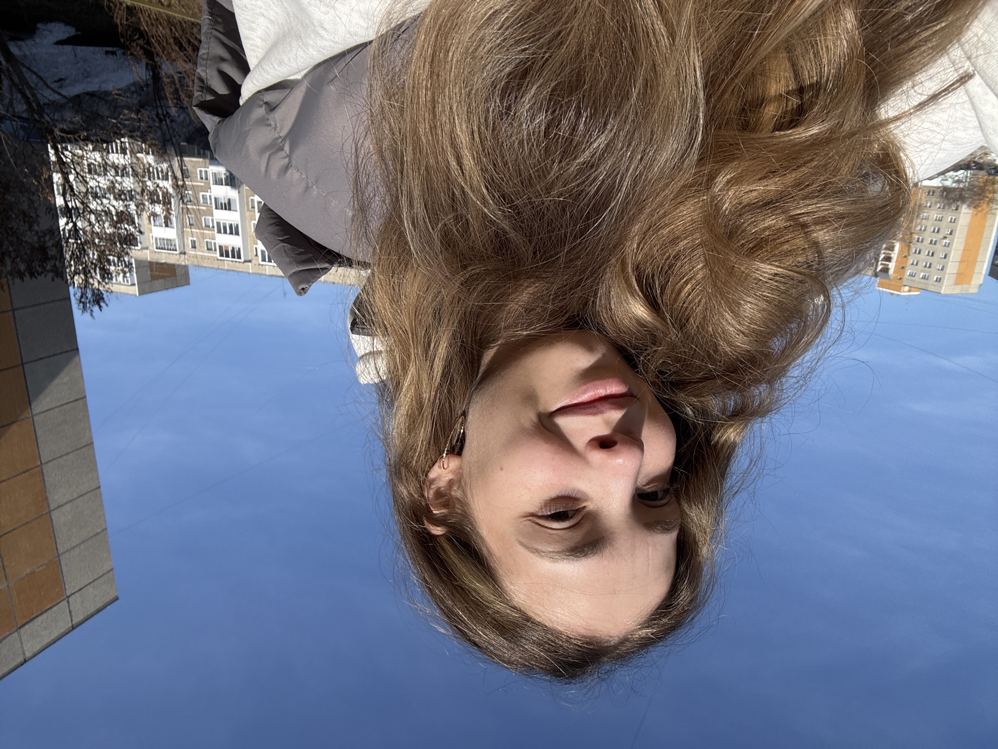 A young woman is pictured in a portrait-style photograph, taken from a low angle and oriented upside down. She has fair skin, light brown hair, and is wearing a jacket. Buildings are visible in the background, suggesting an urban environment. Based on the buildings and general appearance, the location appears to be in the city of Chaikovsky, Russia. The sky is clear and blue.