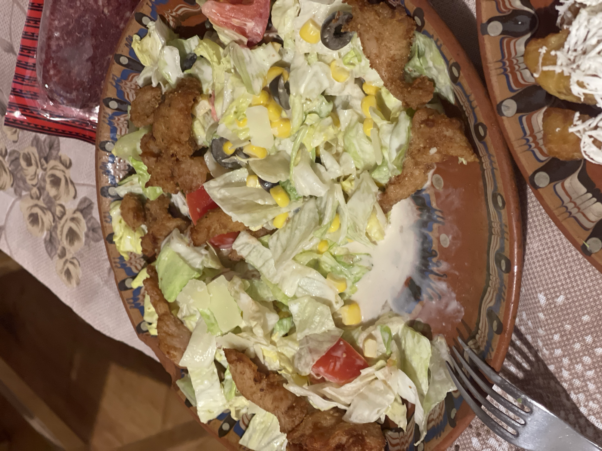 An overhead shot displays a rustic meal laid out on a patterned tablecloth, with two ceramic plates dominating the frame. The central item is a large salad served in a decorative, brown ceramic plate with traditional patterns in blue, white, and black. The salad consists primarily of chopped iceberg lettuce, yellow corn kernels, sliced black olives, diced red tomatoes or bell peppers, and white, likely boiled potato pieces, all generously coated in a creamy white dressing. Scattered throughout the salad are several irregular, golden-brown fried pieces, resembling breaded chicken or fish.
To the upper left of the salad plate, partially visible on the tablecloth, is a packaged item, likely a cured sausage or salami, dark reddish-brown in color and enclosed in clear plastic, resting on a small red and white striped label or packaging.
To the right, another similar ceramic plate is partially visible, holding several golden-fried items, possibly croquettes or polenta pieces, heavily topped with a generous amount of grated white cheese or a similar crumbly ingredient. A silver-colored fork rests on the tablecloth, extending into the lower right portion of the main salad plate.
The tablecloth itself features two distinct patterns: a soft floral design with light-colored roses on a cream background is visible on the left, while a grid of small, light-colored dots or hearts on a darker background covers the right side. The overall setting suggests an indoor mealtime. No people are visible, and no discernible text is present on any items.