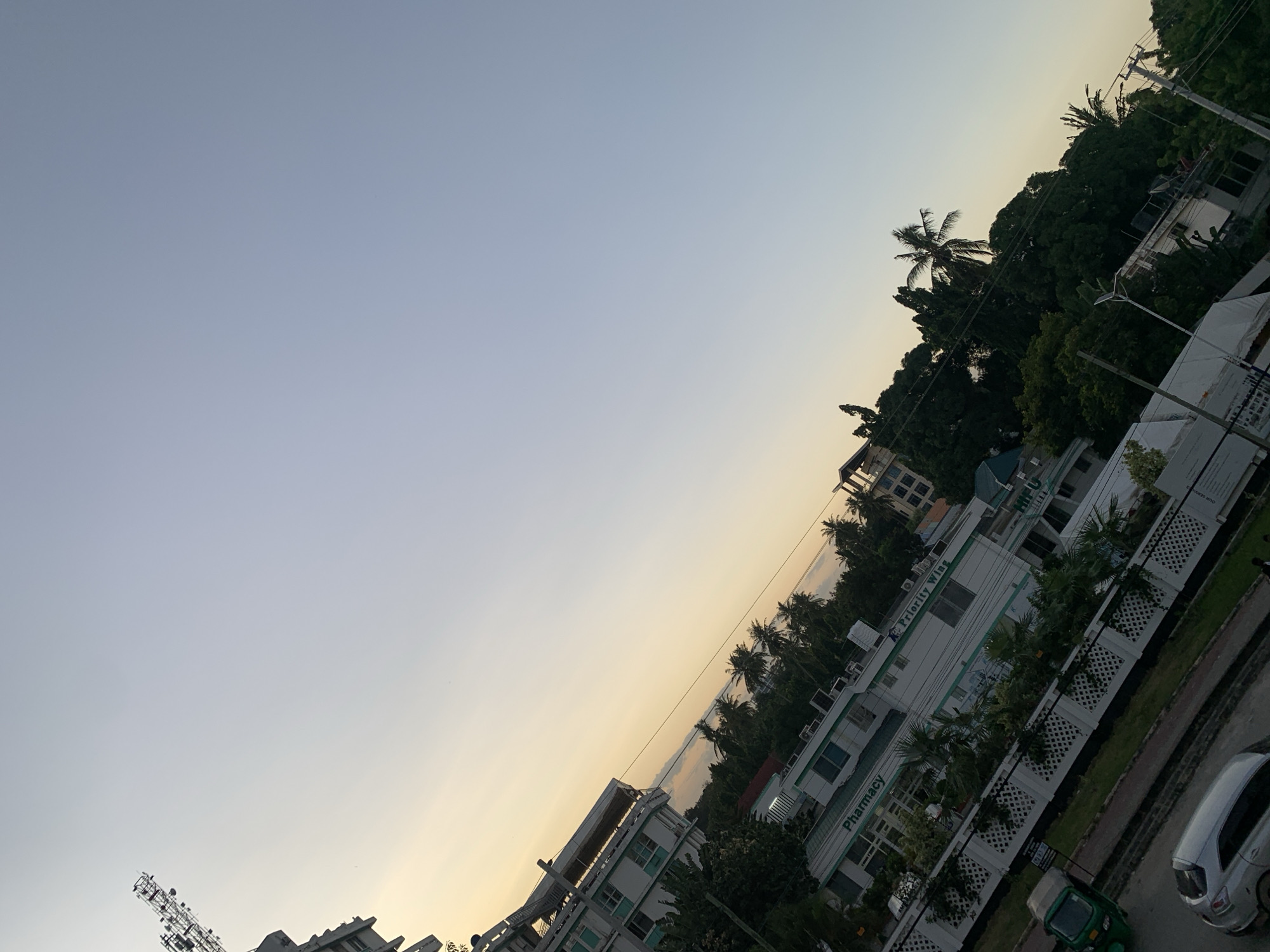 The image, likely taken in Dar es-Salaam, Tanzania, features an oblique view of a cityscape at either sunrise or sunset, indicated by the gradient of light from pale blue to yellow in the sky. The foreground shows the rooftops and upper stories of several buildings, some marked with "Pharmacy" and "Priority Wing," suggesting a medical or commercial district. Palm trees and other greenery punctuate the urban landscape. The background reveals a strip of water, presumably the Indian Ocean, framed by more buildings and vegetation along the coastline. Street traffic is visible, including a silver car and a green tuk-tuk. A communications tower is discernible in the bottom left corner of the image.