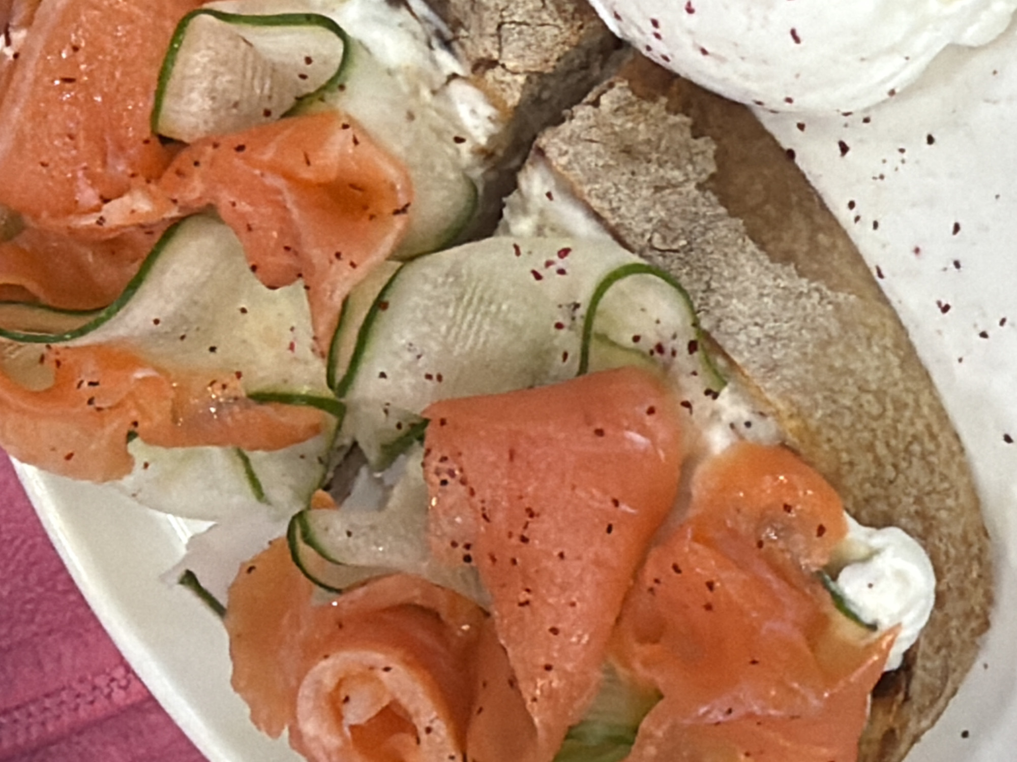 This close-up image displays a gourmet open-faced sandwich or bruschetta. The primary subjects are various food items: vibrant orange-pink slices of smoked salmon, thinly cut cucumbers with green edges, a substantial layer of white creamy spread (likely cream cheese), and a piece of rustic, crusty bread. Small dark red or black specks, resembling ground pepper or paprika, are sprinkled generously over the salmon, cucumbers, and creamy spread.
The salmon slices are artfully arranged, some curled into rosettes, while the cucumbers are almost translucent, suggesting they were peeled or sliced very thinly. The bread, forming the base of the dish, appears to be a hearty, possibly whole-grain or sourdough type.
The dish is presented on a white plate, visible along the bottom and left edges of the frame. A textured pink or magenta textile, possibly a napkin or tablecloth, is visible in the bottom-left corner, adding a soft color accent.
The activity depicted is the presentation of a carefully prepared meal, suitable for breakfast, brunch, or a light appetizer. The lighting is bright and clear, highlighting the freshness and textures of the ingredients. There is no visible text within the image. While the location context of Izhevsk, Russia was provided, the image itself does not contain any visual cues to confirm this geographical detail.