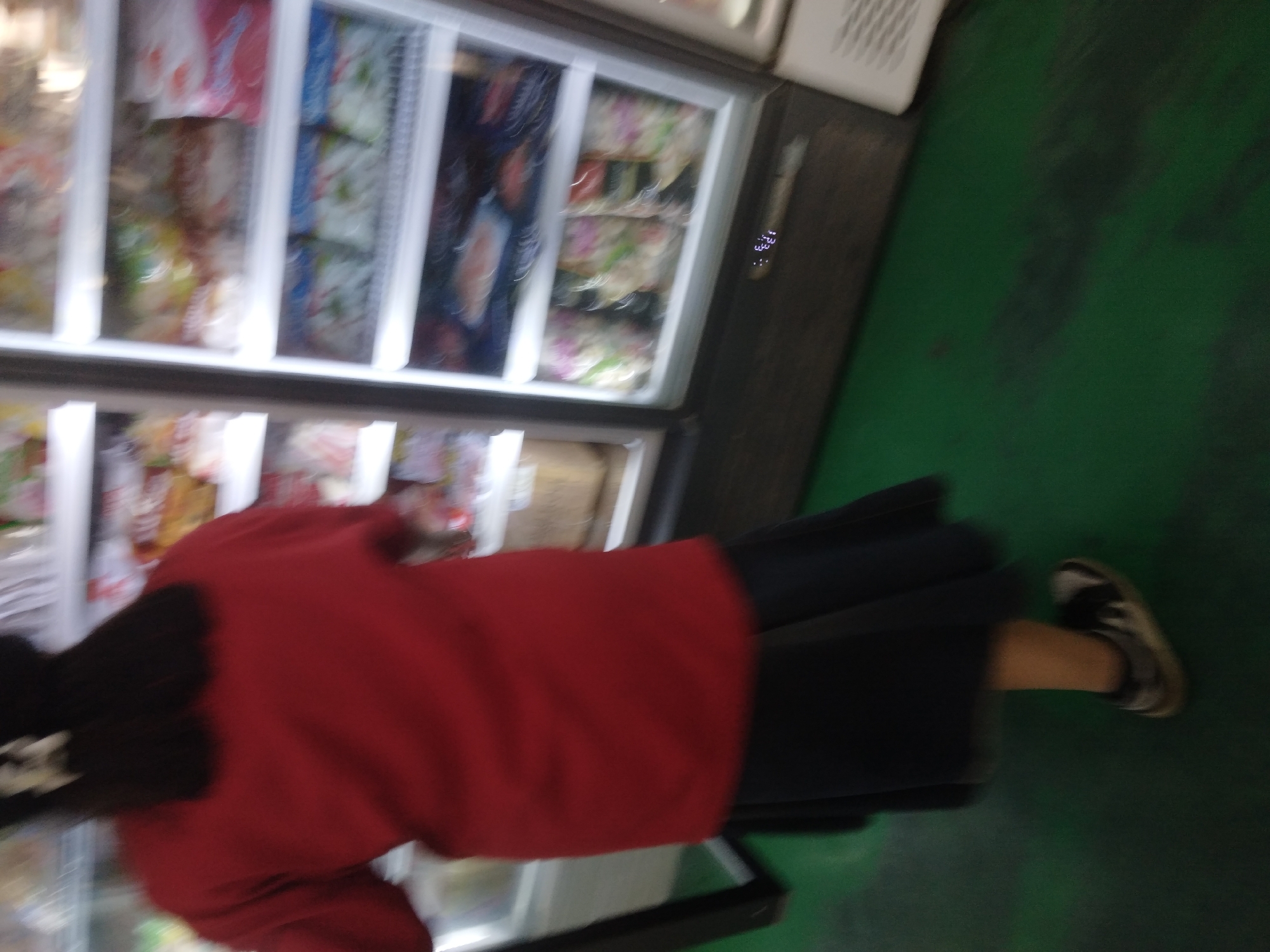 A person wearing a red long-sleeved shirt and a dark blue skirt stands in front of a refrigerated display case filled with packaged food items. The display case is open, revealing multiple shelves with various products. The person's back is to the camera, and they are looking into the display. The floor is a dark green color, and the scene appears to be indoors, possibly in a convenience store or market in Sokaraja, Indonesia. A white bow is visible in the person's dark hair.