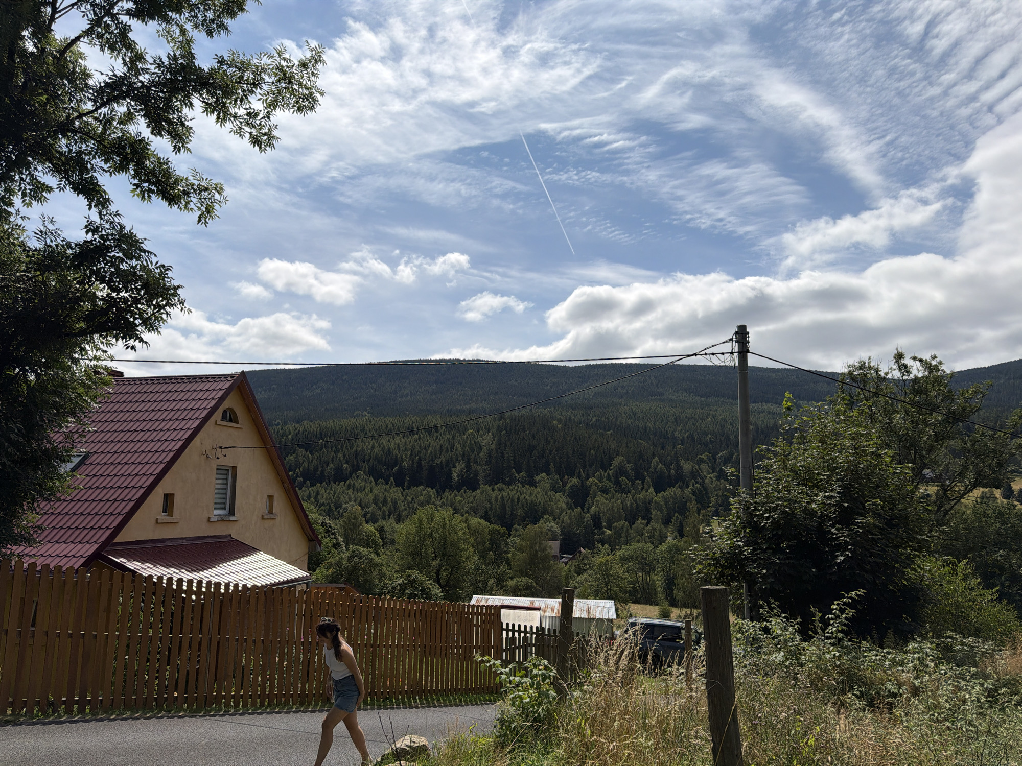 An outdoor, medium shot captures a woman walking on a paved road adjacent to a wooden fence. The woman, positioned in the lower left of the frame, wears a white tank top and denim shorts, with her body angled away from the camera. Behind the fence, a yellow house with a red tiled roof is visible. The background features a dense forest that extends to a distant, tree-covered mountain range under a sky with scattered clouds and a faint airplane contrail. The scene is set in Świeradów-Zdrój, Poland, suggesting a rural or semi-rural environment with a clear day.
