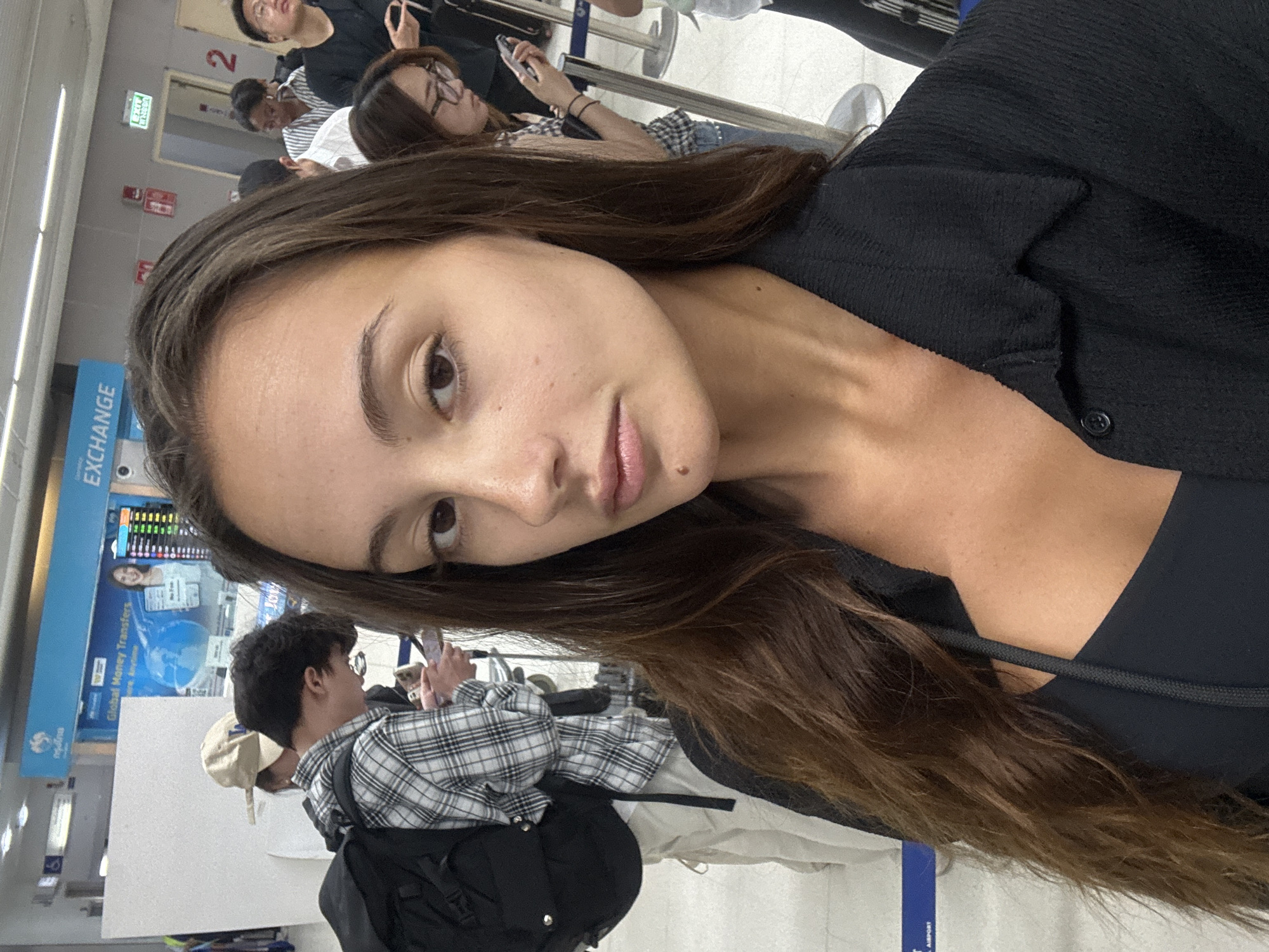 A young woman with long, dark brown hair and a neutral expression takes a selfie, dominating the foreground. She wears a black long-sleeved top over a dark bra or camisole. A small mole is visible on her chin. The image is taken in an indoor public space, likely an airport or a travel terminal, indicated by queue barriers and signage in the background.
Several other people are visible behind her, appearing to be waiting in line. One person directly behind her wears a black and white plaid shirt and a backpack, looking at a smartphone. Further back, another individual with glasses is also focused on their phone.
The background features a blue information board with the word "EXCHANGE" prominently displayed, alongside "Global Money Transfers" and a graphic of a globe. An illuminated screen below shows various currency exchange rates. On the wall, a green "EXIT" sign, a red fire alarm pull station, and a large number "2" are visible. The flooring consists of light-colored tiles. The setting suggests a busy travel hub, consistent with an airport in Tambon Mai Khao, Thailand.