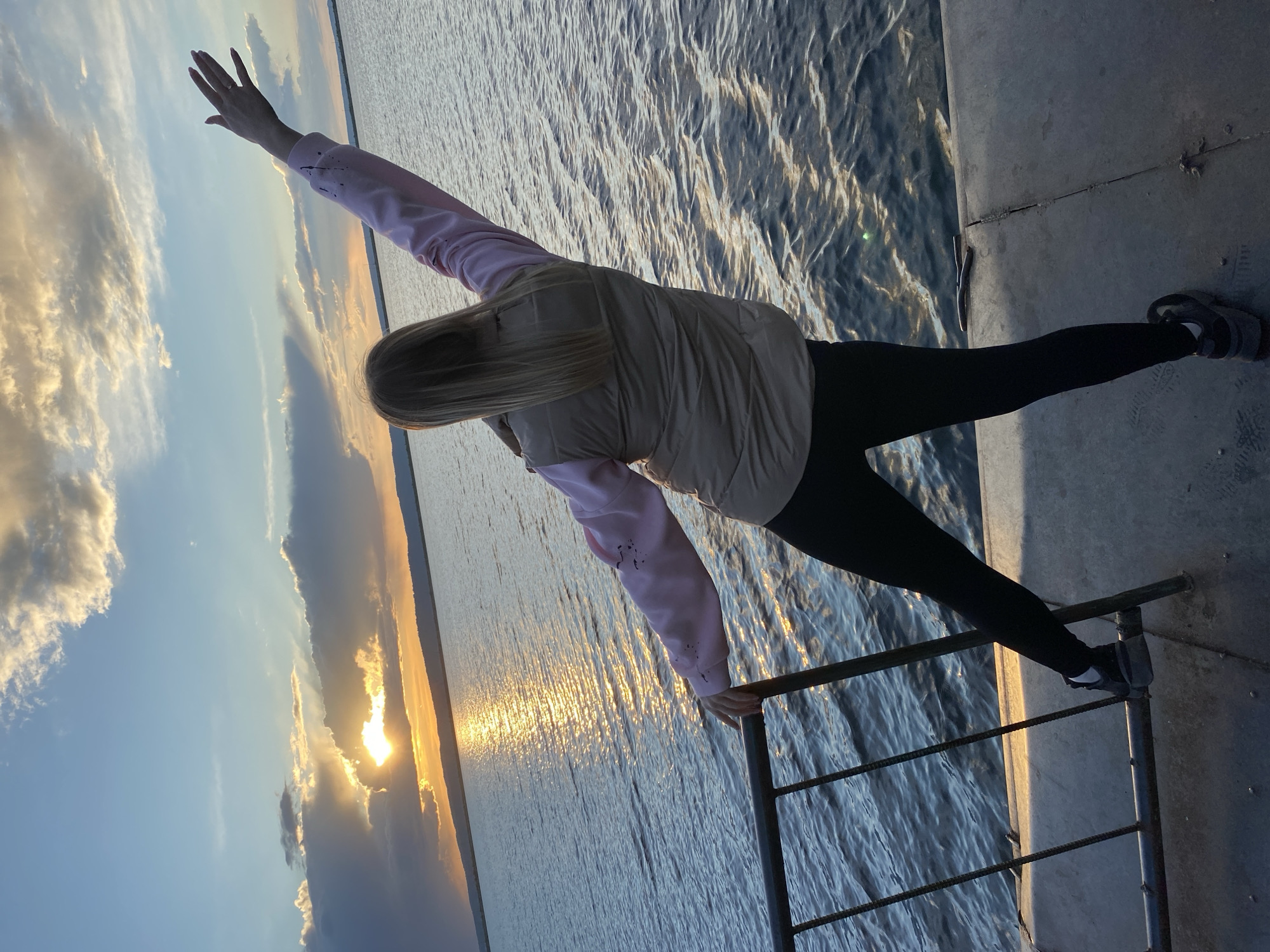 A woman performs a balancing act on a railing near a body of water at sunset. She is wearing a light pink and beige jacket, black pants, and sneakers. Her left hand grips the railing for support, while her right arm extends upwards toward the sky. Her right leg is straight and extends out to the side, while her left leg remains straight and connects her to the railing. The background features a body of water reflecting the light of the setting sun. Clouds are visible in the sky, adding texture to the scene. The location is likely the city of Chaikovsky, Russia, based on the provided context.