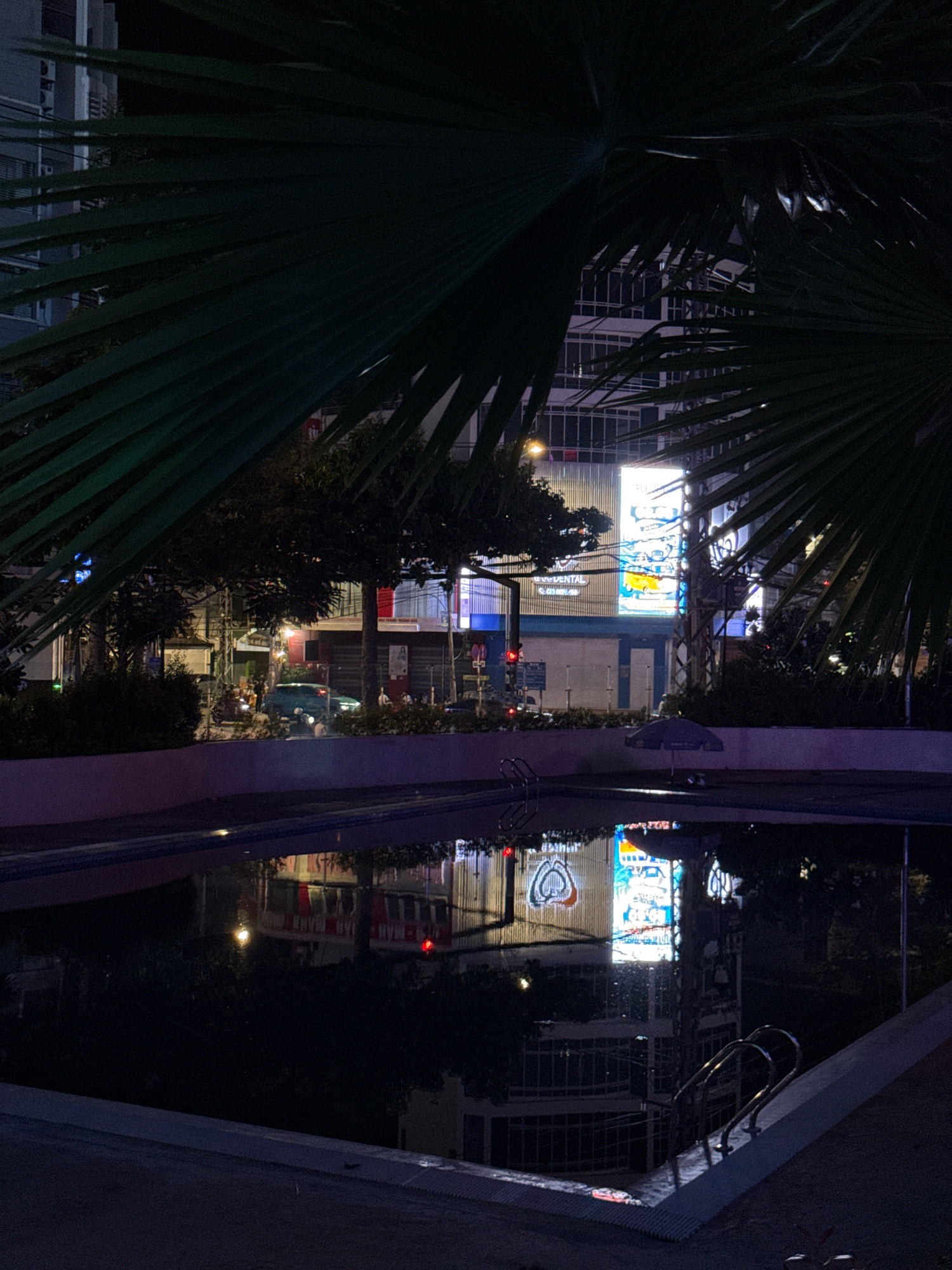 This is a nighttime exterior shot of a swimming pool in Nha Trang, Vietnam. The pool's water reflects the surrounding urban environment, including buildings illuminated by bright neon signs and streetlights. Large palm fronds in the foreground cast shadows, partially obscuring the view. In the middle ground, a dark-colored umbrella stands near the pool's edge. The background features multi-story buildings with numerous windows, some lit up, and a prominent billboard displaying colorful advertisements. A few vehicles and motorcycles are visible on the street, suggesting a moderate level of activity. The overall atmosphere is dark and moody, characteristic of a tropical city at night.