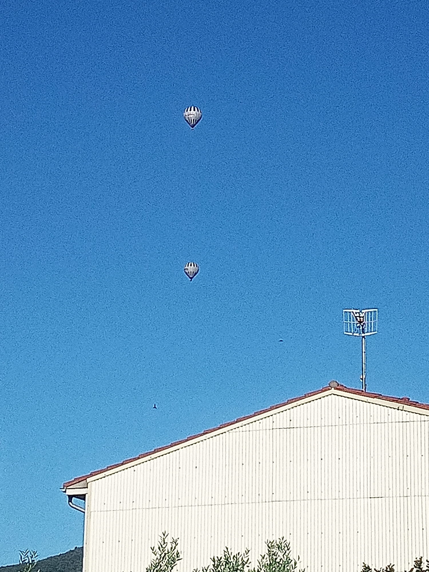 The image captures a clear blue sky over Sant Joan Les Fonts, Spain. Two hot air balloons, one higher and to the left, the other lower and to the right, are visible against the sky. Both balloons have a distinct black and white striped pattern. In the foreground, the white corrugated siding of a building with a red-tiled roof is visible. A metal television antenna with a square grid on top is mounted on the roof. A dark shape, possibly a bird, is present in the sky to the left of the antenna. There is also a smaller dark shape in the sky to the right of the house, just above the roofline. The lower part of the image shows greenery, including some trees and shrubs. The overall scene suggests an outdoor view, possibly from a residential area.