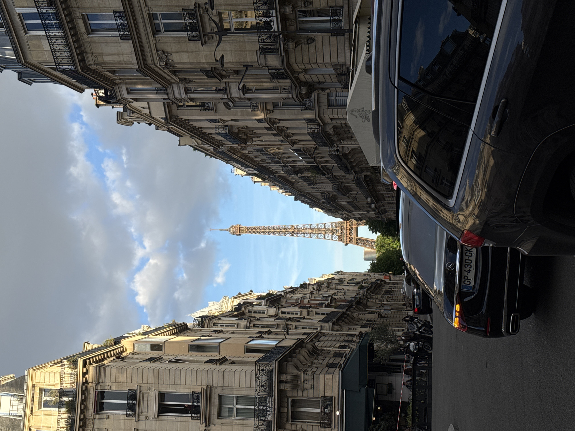 The image displays a classic Parisian street scene, looking down a relatively narrow thoroughfare towards the iconic Eiffel Tower in the distance. Tall, ornate Haussmannian-style buildings, characterized by their light stone facades, numerous windows with decorative iron railings, and multiple stories, line both sides of the street, converging as they recede into the background.
On the left, the buildings showcase intricate architectural details and several balconies. The sky above transitions from patches of clear blue with scattered white clouds closer to the horizon to a larger mass of darker, grey clouds on the upper left. Some greenery is visible on the rooftops of certain buildings, and in the foreground on the left.
On the right, a dark-colored SUV or large sedan is parked prominently in the foreground, with its rear facing the viewer. The car's tinted windows reflect the architecture of the buildings opposite. A European license plate is visible on the rear, displaying the characters "GP-430-GK" along with the "F" country code. Further down the street on the right, additional vehicles, including another car and several motorcycles, are parked. Below the buildings on this side, there's a glimpse of what appears to be a storefront or cafe with an awning.
The Eiffel Tower stands centrally in the mid-distance, its lattice structure clearly visible against a relatively bright blue sky with some soft clouds. The lighting suggests daytime, possibly late afternoon, with a mix of direct sunlight illuminating the tower and parts of the buildings, while other areas are under the shadow of passing clouds. The scene captures the grandeur of Parisian architecture leading to one of the world's most recognizable landmarks.