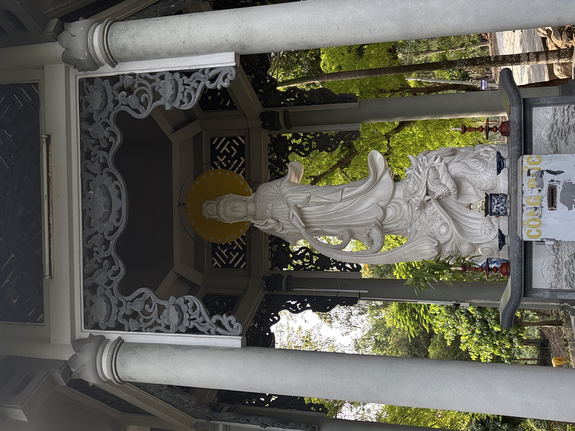 The image shows a white statue of a woman standing on a dragon within a temple-like structure. The statue is adorned with a yellow halo and intricate carvings. The structure is supported by large, grey columns and features detailed stonework. In front of the statue, there is an altar with smaller figurines and decorative items. The background consists of lush green trees and foliage, suggesting an outdoor setting. The text on a plaque in front of the altar is partially visible and appears to be in Vietnamese. The overall scene depicts a religious or spiritual monument in a serene, natural environment. This monument is located in Tay Nha Trang Ward, Vietnam.