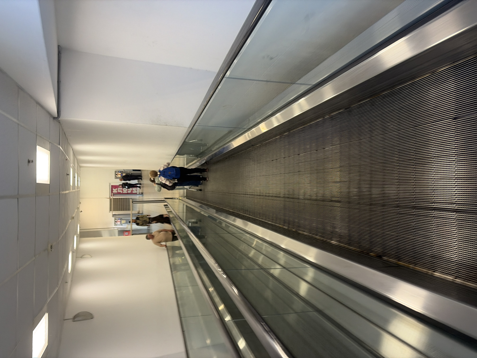 An indoor scene depicts a long, well-lit passageway featuring a moving walkway, commonly known as a travelator. The image, initially oriented vertically, shows the travelator dominating the right side of the frame, extending far into the distance. Its surface is composed of textured, dark metallic strips, flanked by shiny silver-colored metal borders and clear glass panels, topped with a metallic handrail.
Several individuals are present. In the mid-ground, a person wearing a blue backpack and a light-colored, striped top stands on the travelator, facing away from the viewer. Further along the travelator, another person in a light beige top and dark bottoms is also visible. In the background, to the left of the travelator, a small group of people are standing near what appears to be a storefront or an entrance.
The surrounding environment consists of smooth, white walls and a white suspended ceiling with square panels. Rectangular recessed lights are spaced along the ceiling, providing bright artificial illumination. Additionally, several semi-circular, wall-mounted lights are visible on the left wall, complementing the overhead lighting.
The setting suggests a modern commercial space, such as a shopping mall or an airport terminal. Activity is limited to people riding or standing near the moving walkway. No specific time of day is discernible, but the bright artificial lighting indicates an indoor setting independent of natural light.
A notable detail is a red sign with white text visible in the background, which appears to be in Cyrillic script. The characters resemble "УЮТ", which means "comfort" or "cozy" in Russian, aligning with the provided location context of Sochi, Russia.
